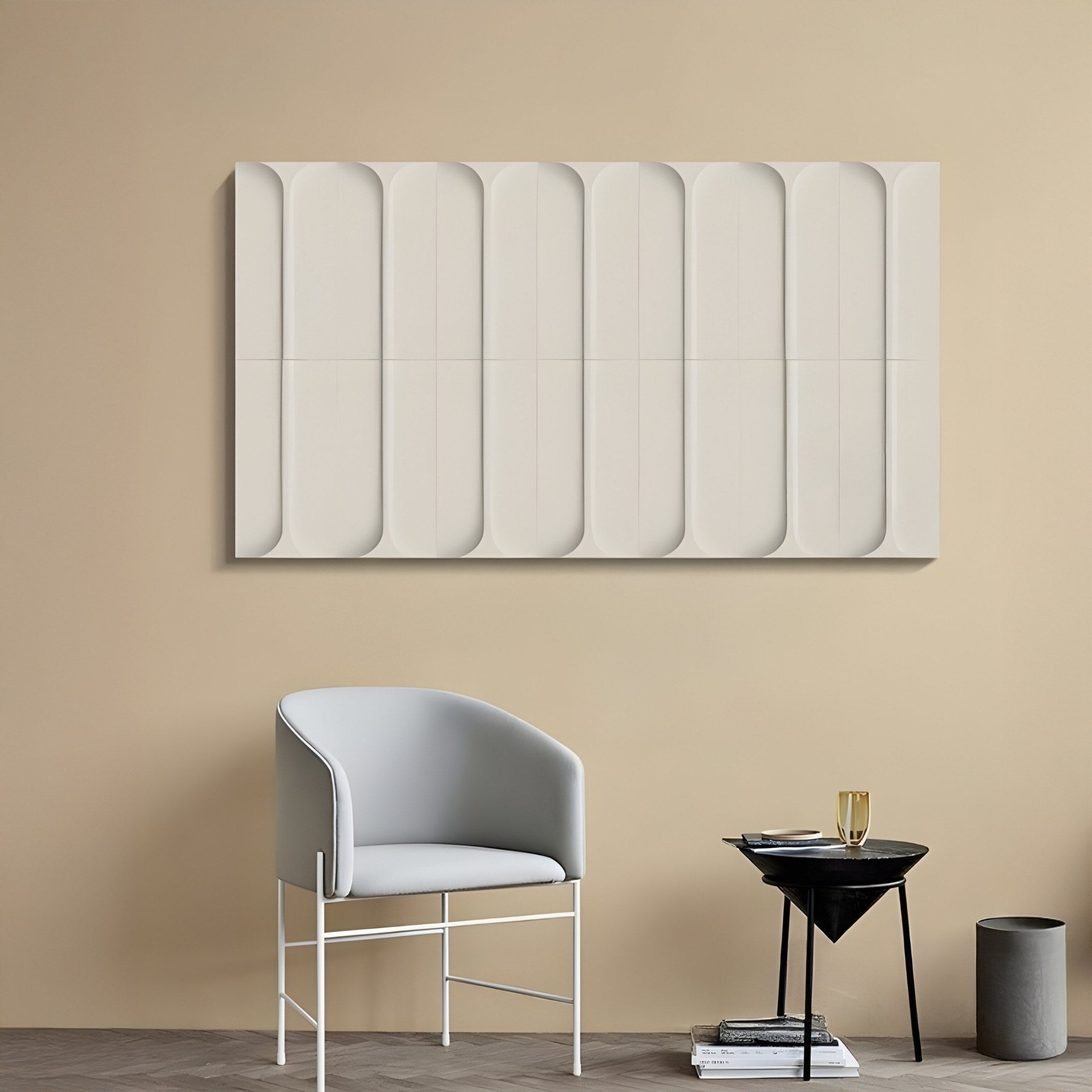A minimalist room with a gray chair and black triangular side table is enhanced by the Vireus White Short Dual Panel 3D Wood Relief Wall Art from Giant Sculptures. A gold-trimmed glass with a drink and stacked books sits on the table. Light wood floors and beige walls complete this elegant decor.