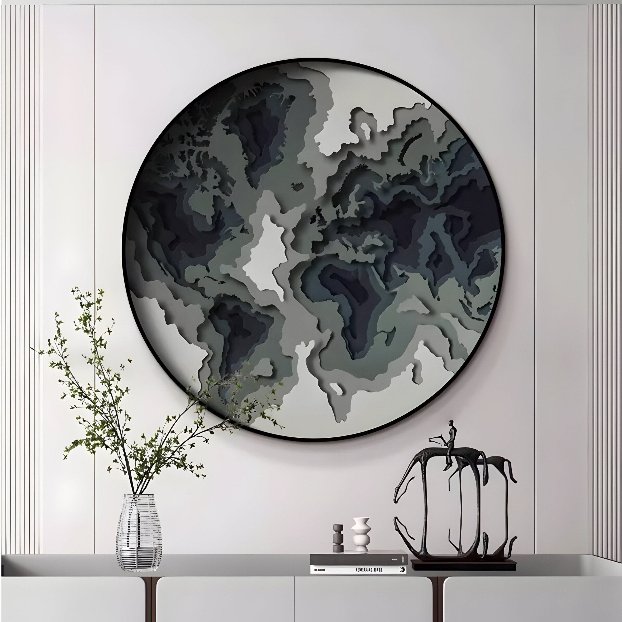 A modern interior features the Terranova Earth Green Round Wood Carving 3D Wall Art by Giant Sculptures, showcasing layered shades of gray and black. Below, a minimalist shelf displays a clear vase with branches, a small sculpture, and decorative blocks against the light-colored backdrop.