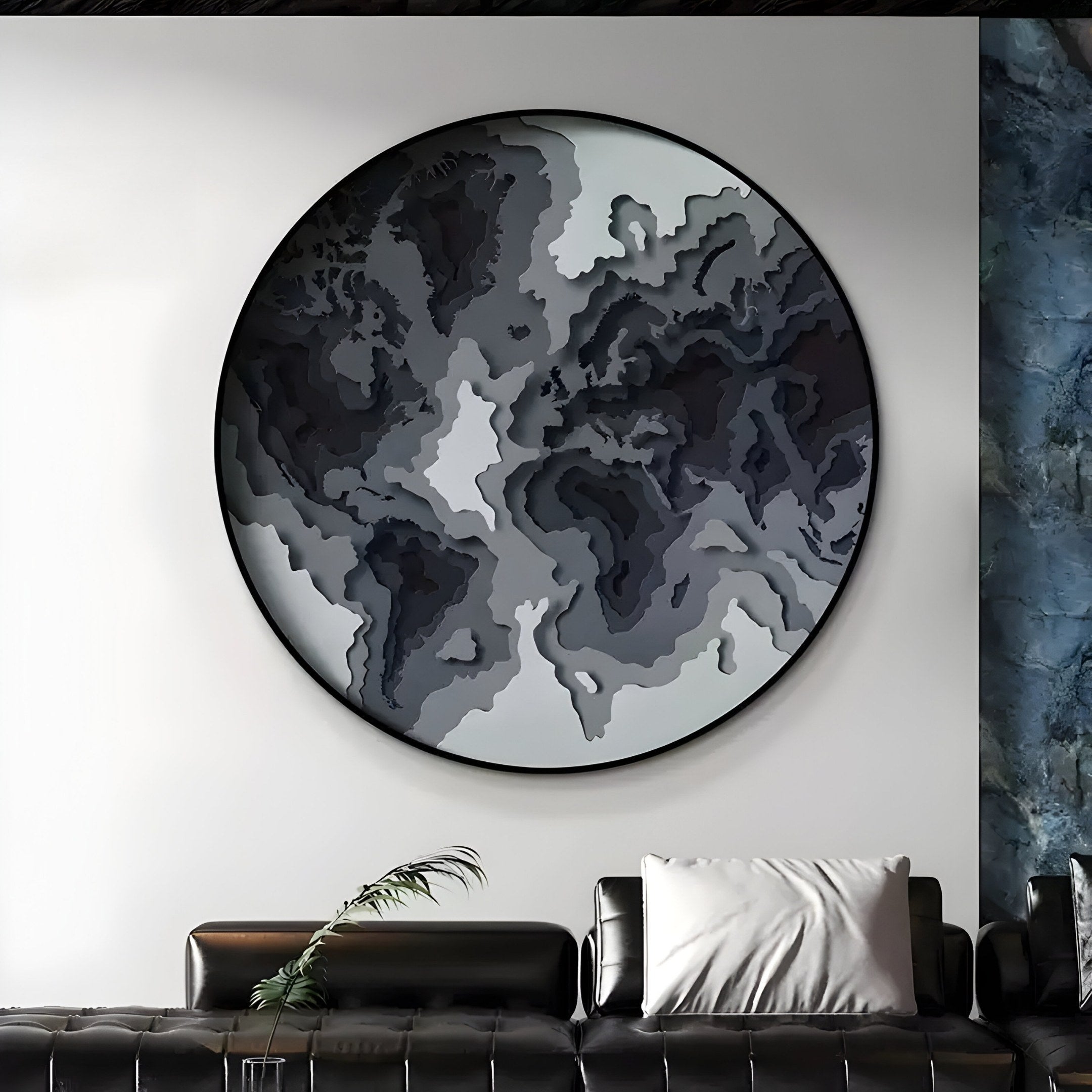 The Terranova Shadow Black Round Wood Carving 3D Wall Art by Giant Sculptures, with intricate gray and white patterns, hangs above a black leather sofa featuring a white pillow. A long leaf on the sofa complements the textured blue and gray wall behind.