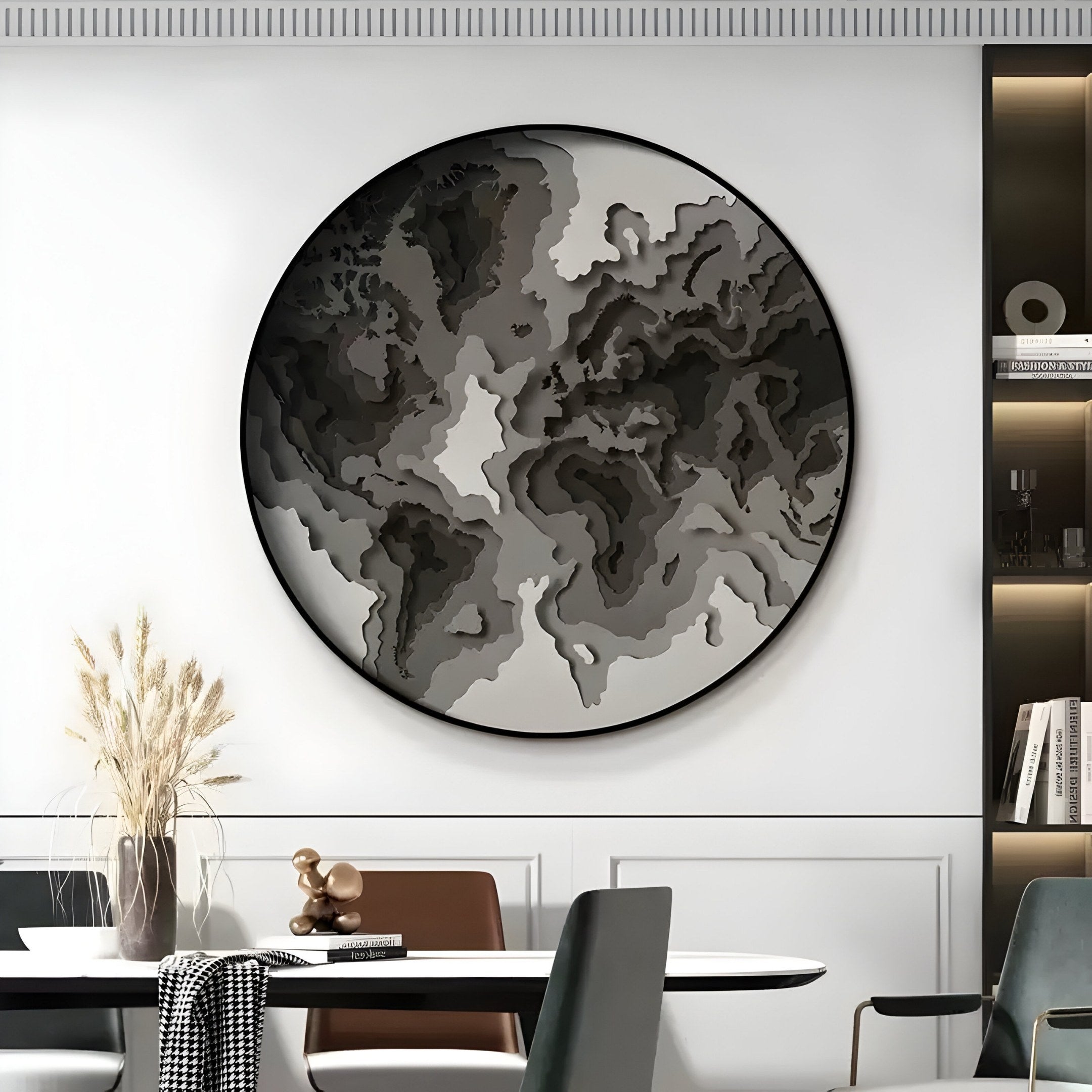 The Terranova Ash Grey Round Wood Carving 3D Wall Art by Giant Sculptures, with its textured sophistication, features an abstract world map. It complements a modern dining table adorned with wheat and nearby bookshelves in black, gray, and white tones.