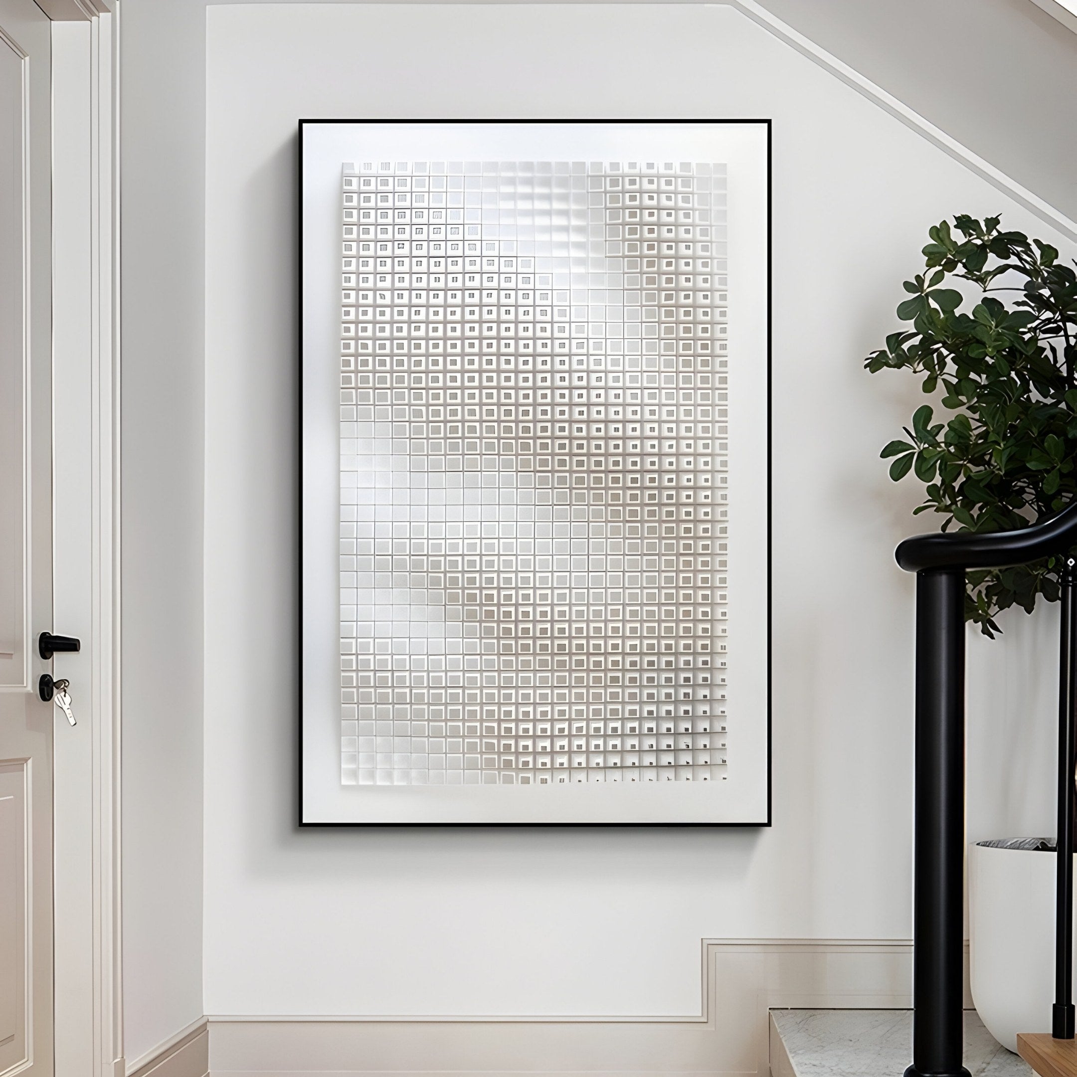 The Elytrum White Grid Hand-Painted Wood Relief 3D Wall Art by Giant Sculptures, featuring a modern geometric design of metallic squares and framed in black, hangs on a hallway wall. It subtly reflects light, while a nearby plant adds life beside the sleek black staircase handrail.