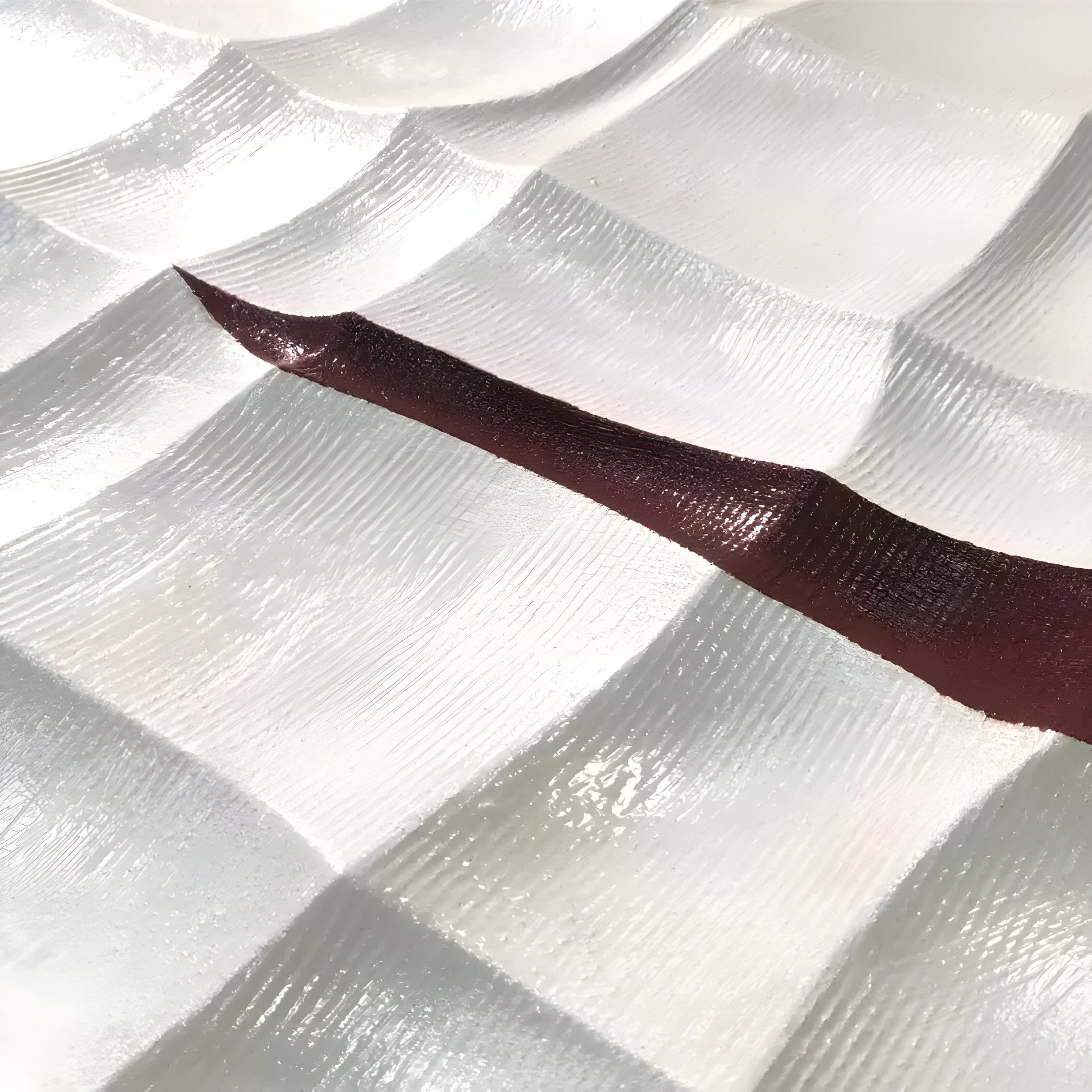 The Striata White & Burgundy Wood Carving 3D Wall Art by Giant Sculptures showcases a textured white surface with wave-like patterns. A shiny, dark burgundy stripe diagonally contrasts the light background as sunlight highlights and casts subtle shadows on the ridges.