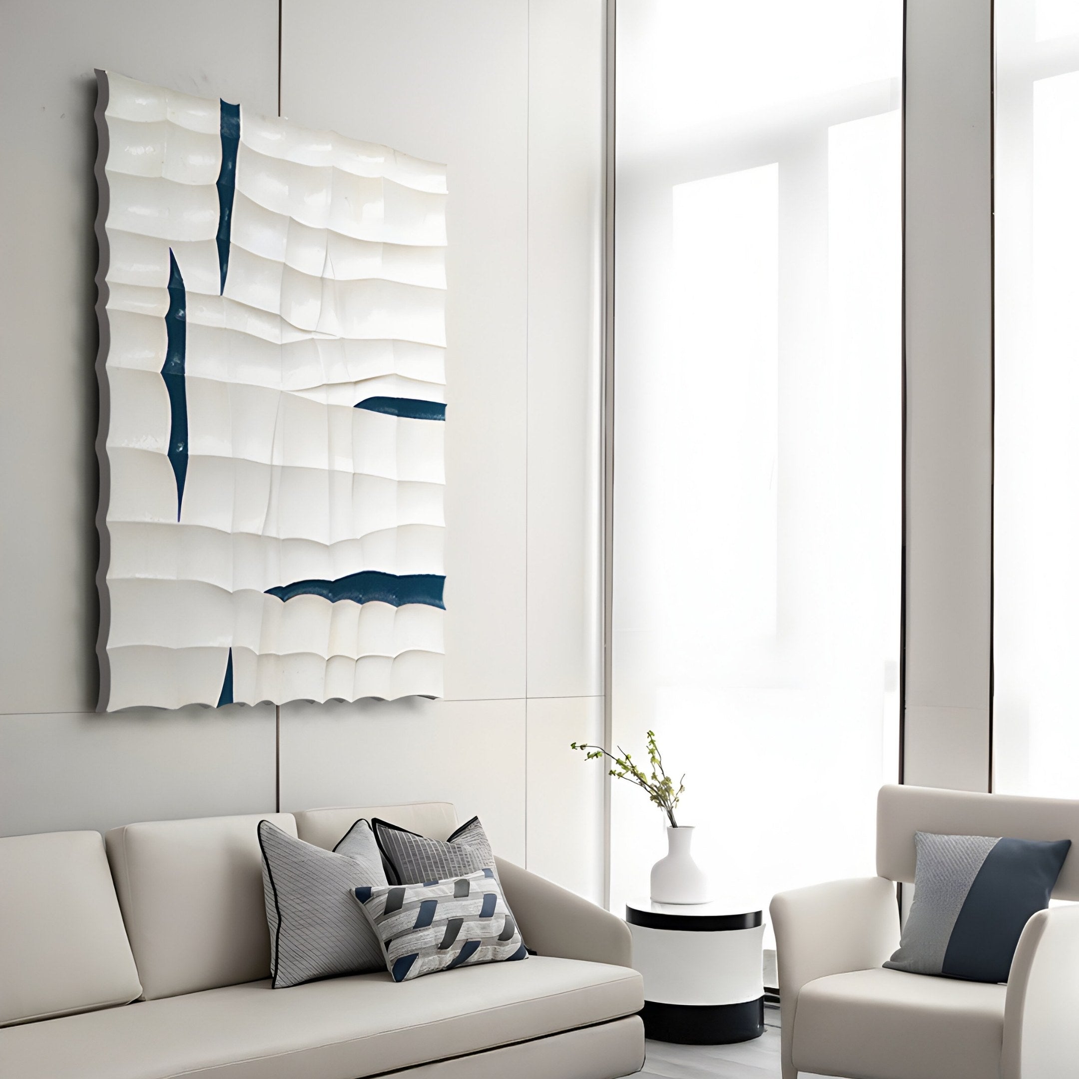 In a modern minimalist living room, a white sofa and armchair with gray cushions and blue accents set the scene. Above, the Striata White & Blue Wood Carving 3D Wall Art by Giant Sculptures adds dimension. A small round table with a white vase sits between the seating.