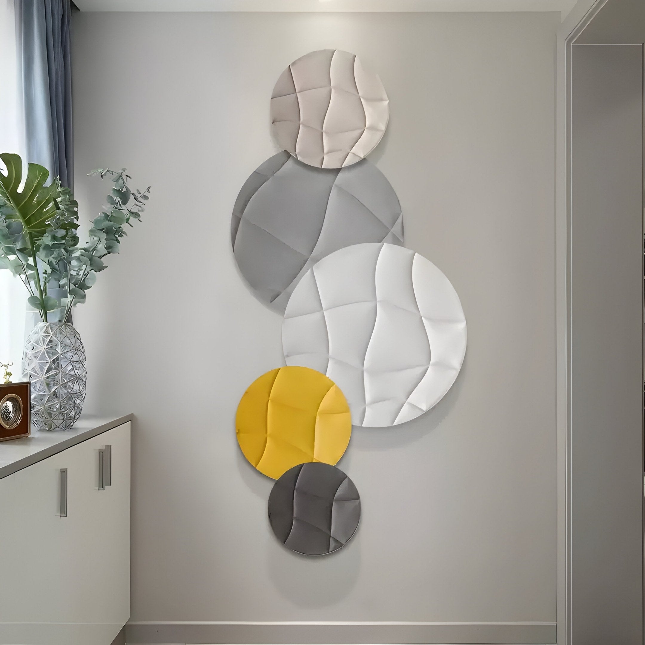 The Giant Sculptures Orbura 5-Set Circular Grey & Yellow 3D Wood Relief Wall Art enhances the light-colored wall with its textured circles. A patterned vase and plant on a nearby cabinet complement the decor, as soft natural light filters through a window.