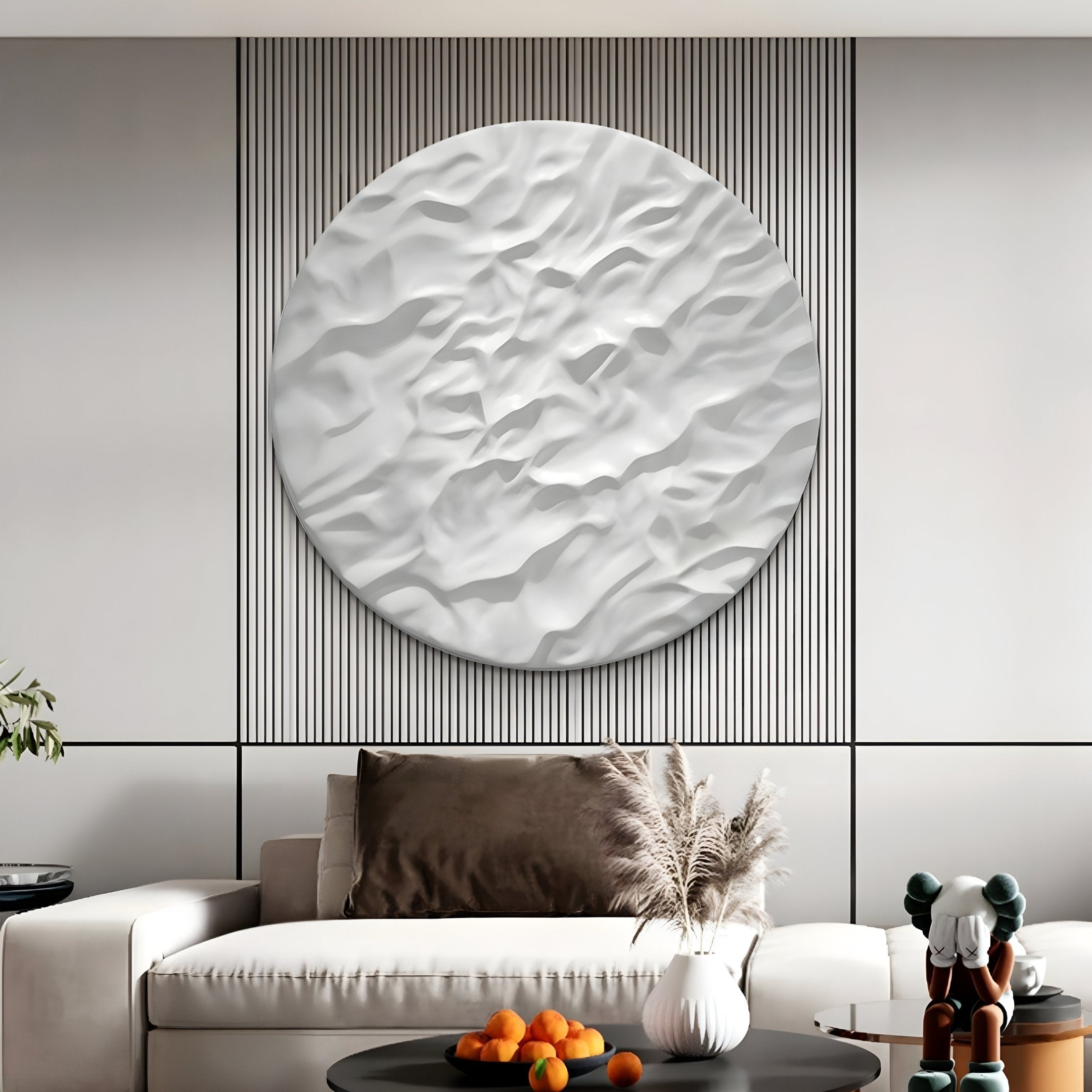 A minimalist living room showcases a beige sofa, decorative grasses in a vase on a black round table, and the Orenda Organic Wave White Wood 3D Wall Art by Giant Sculptures. A small cluster of oranges is on the table, while a sculptural figure stands nearby.