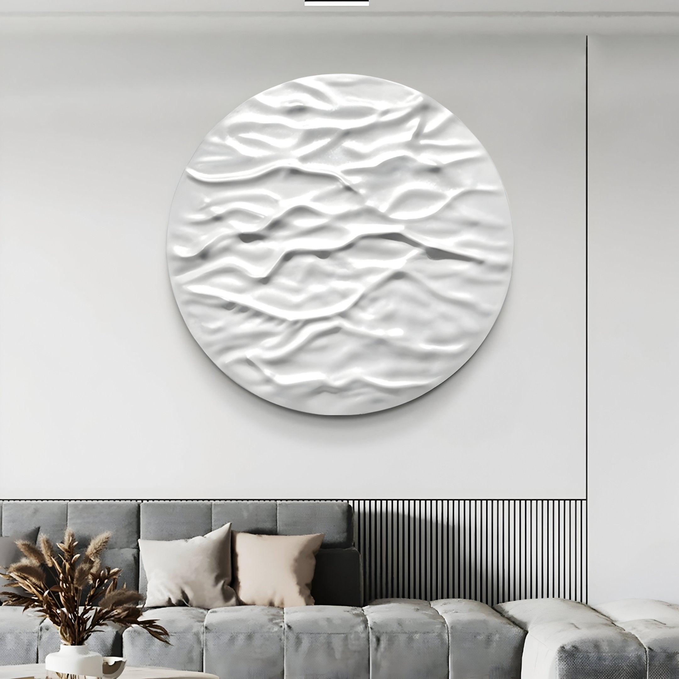 In a modern living room, the Orenda Ripple Wave White Wood Relief 3D Wall Art by Giant Sculptures serves as a striking focal point. Below, a gray sectional is accented with light pillows. A round table holds a decorative vase with dried branches, enhancing the minimalist design.
