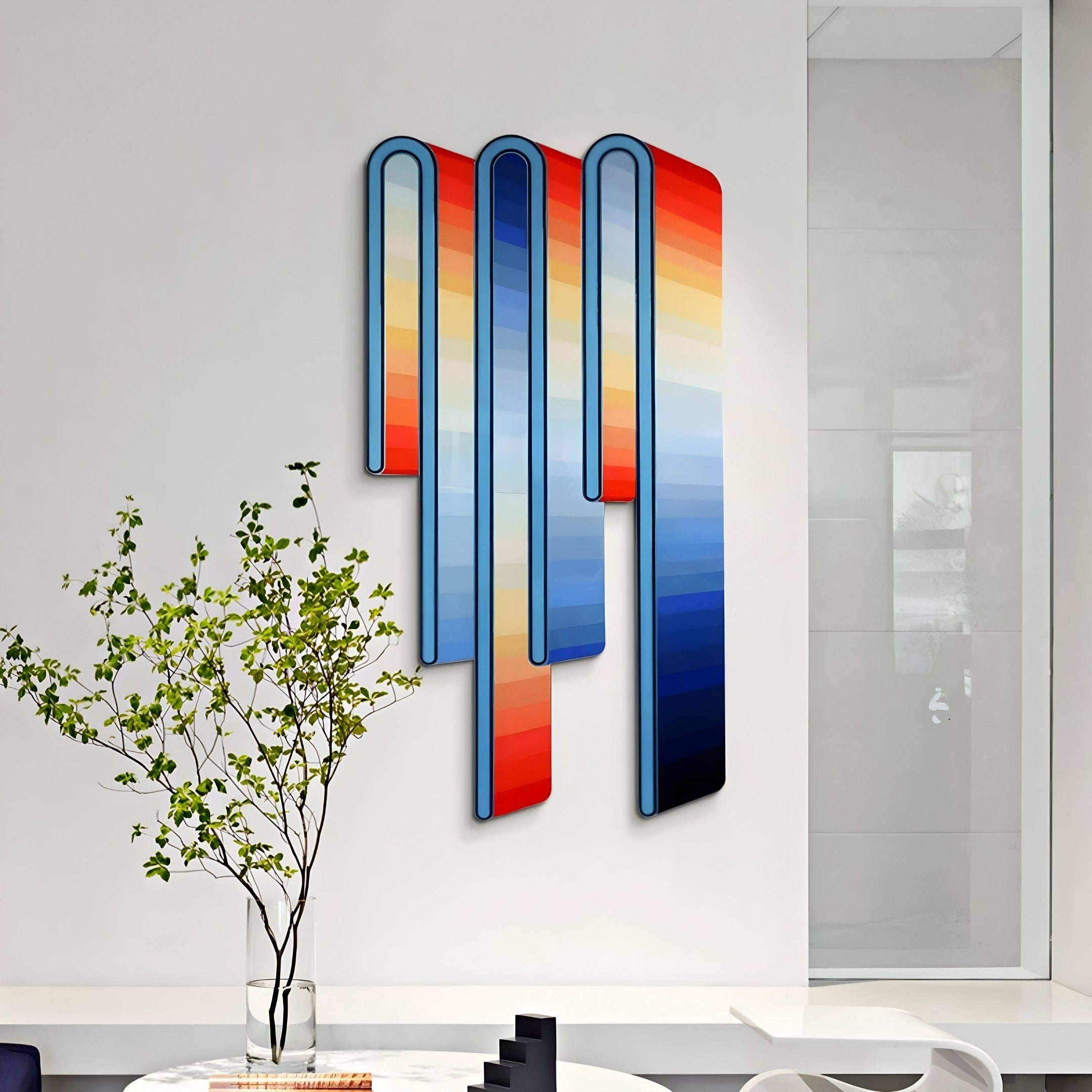 A modern 3D wall art piece, the Opaline Sunset Gradient Wood Relief by Giant Sculptures, showcases elongated vertical shapes with colors shifting from red to blue against a white wall. In this minimalist setting, a potted green plant and black geometric objects rest on a white table.