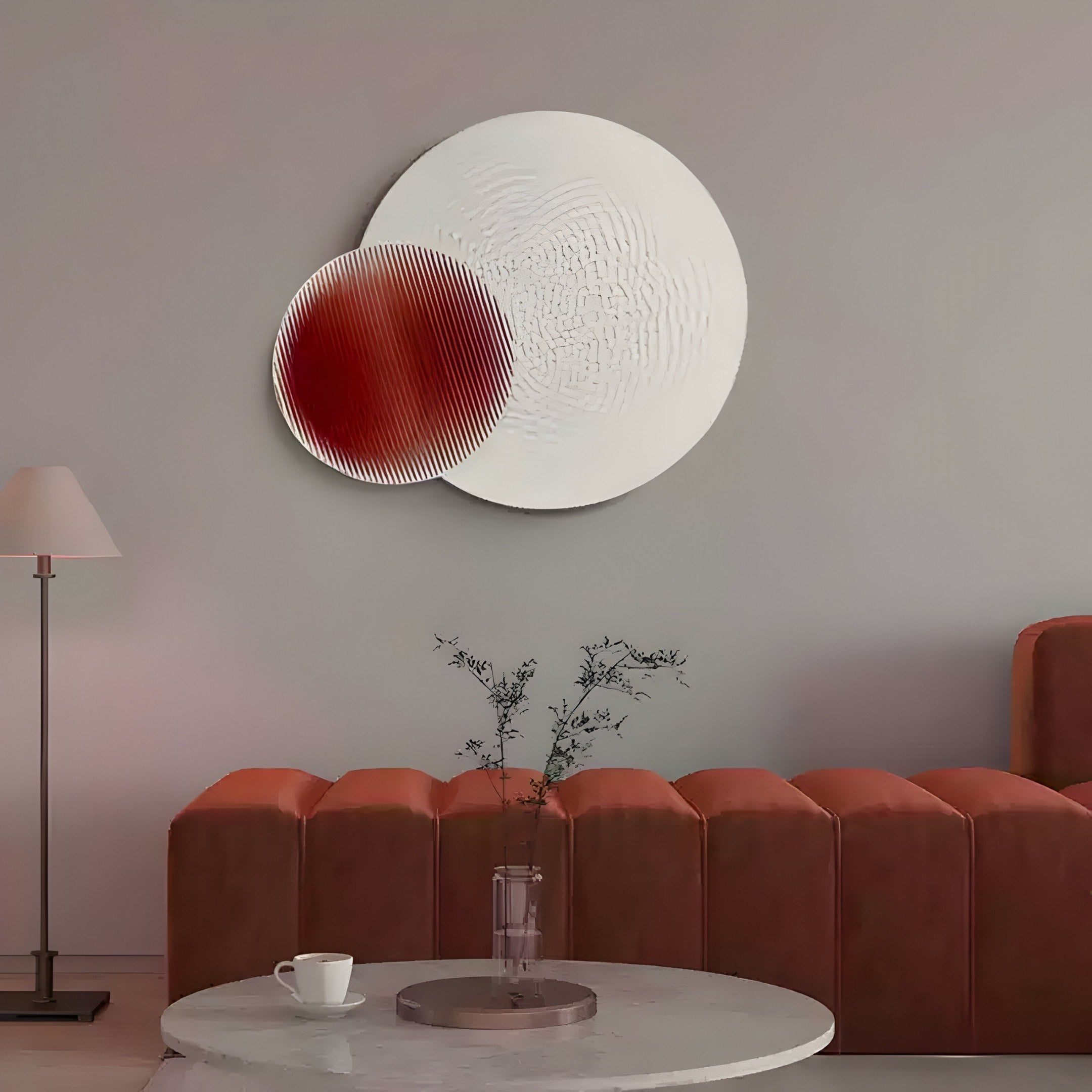 A minimalist living room showcases a round coffee table with a cup and vase of branches. The wall art features the Lucentis White & Burgundy Wood Relief by Giant Sculptures, adding texture. A brown sofa and standing lamp in the background complete the contemporary decor.