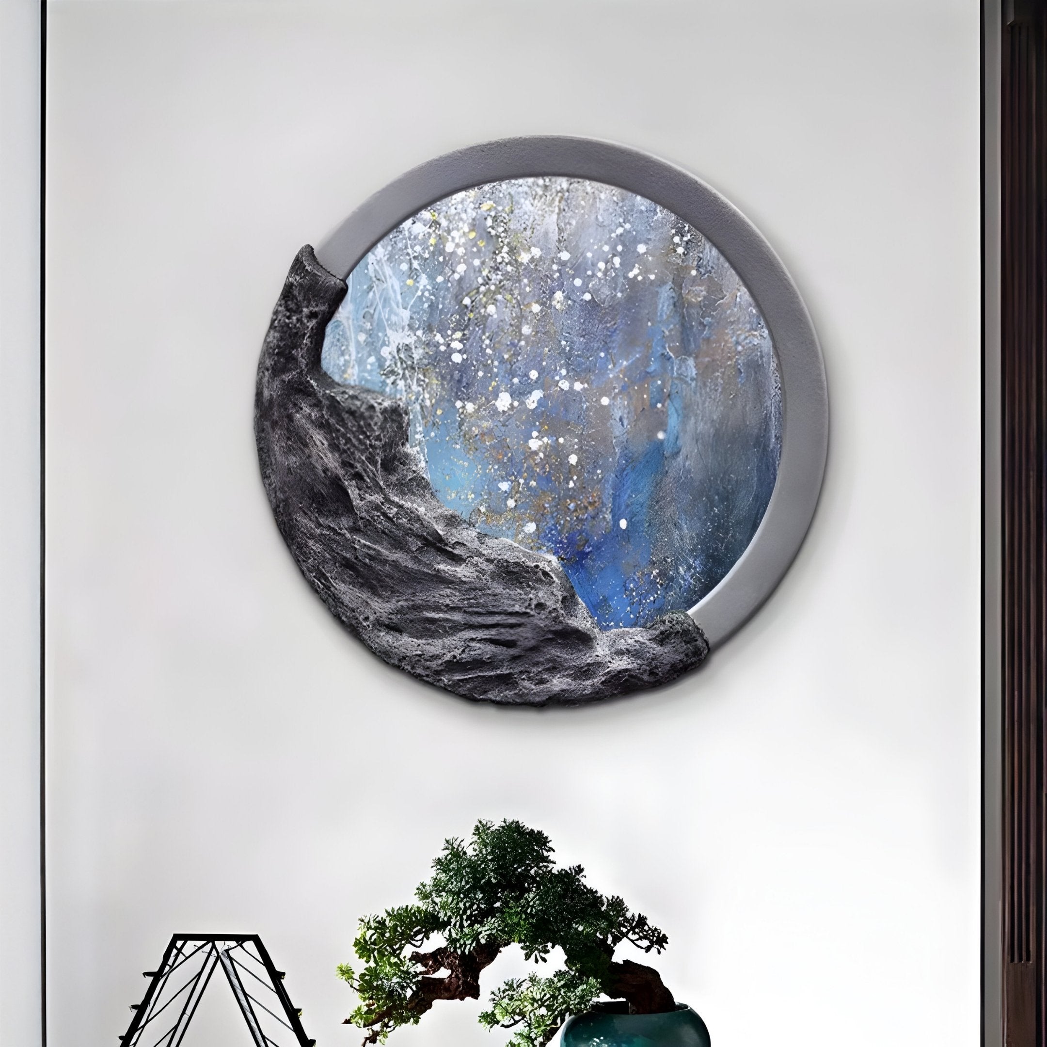 A round mirror with a partial black rock texture frames a starry, misty scene echoing Giant Sculptures Auroria Velvet Dusk Hand-Painted Wood Relief, in blue, gray, and white. Below it rests a lush bonsai tree on a modern geometric stand.