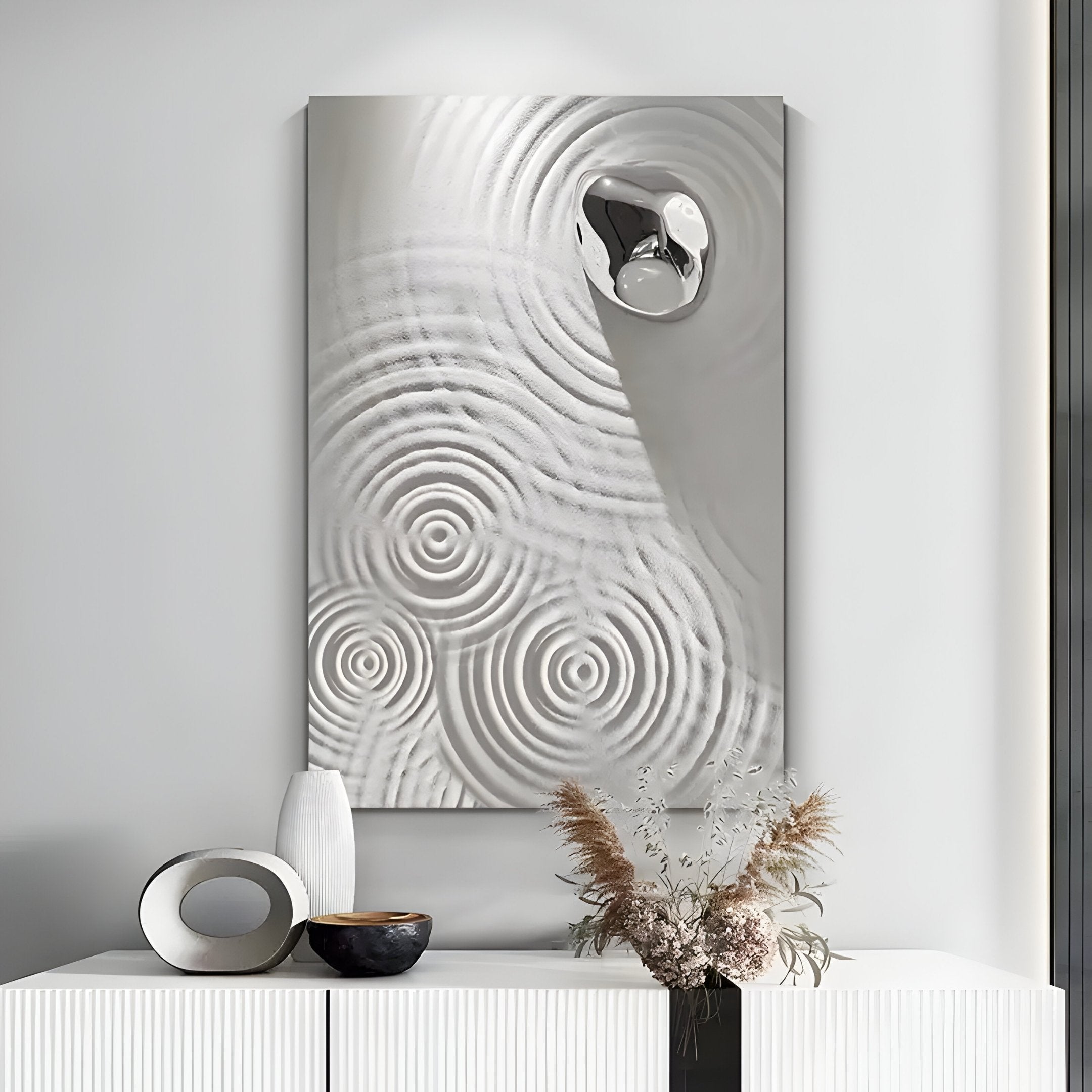 The Ondella Silver Ripple IV Sandstone Wood Relief 3D Wall Art by Giant Sculptures embodies Wabi-Sabi aesthetics with concentric circular patterns and a mounted reflective silver object. Below, a white table showcases decorative vases and dried foliage.