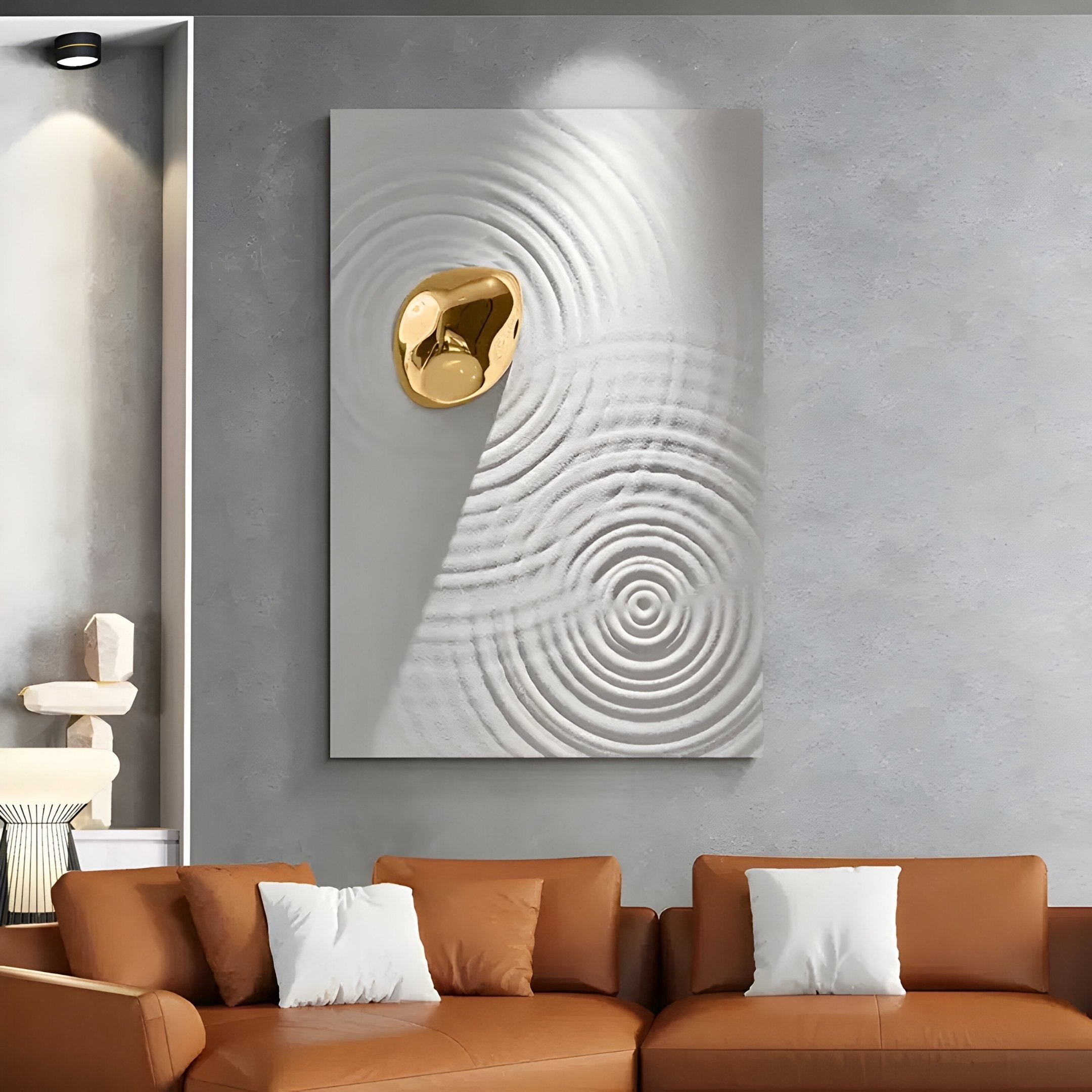 A modern living room features a brown sofa with white cushions. The wall is highlighted by the Ondella Gold Ripple I Sandstone Wood Relief 3D Wall Art by Giant Sculptures, set against concentric circles on a textured gray backdrop. A niche sculpture and soft lighting complete the ambiance.