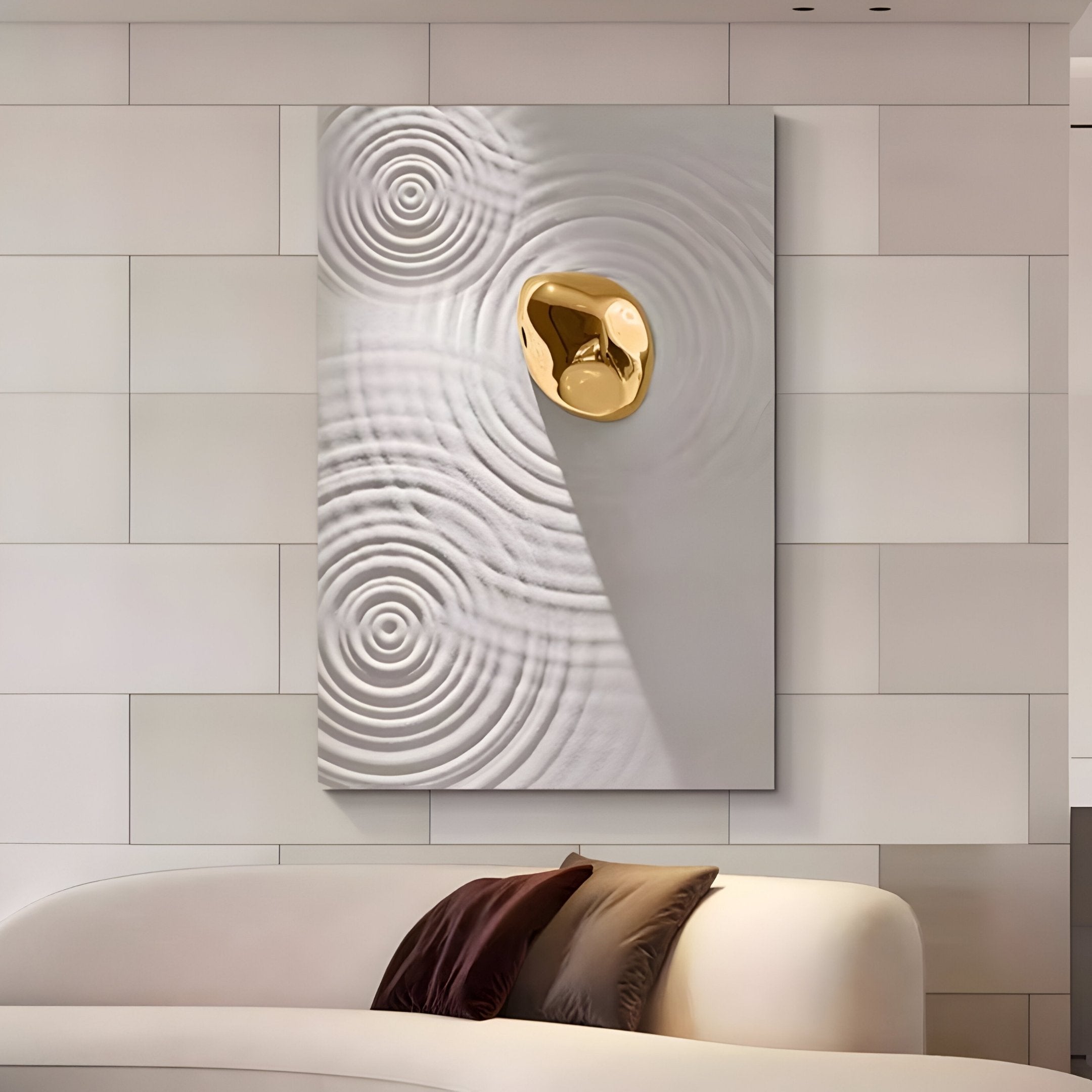 A modern living room features the Ondella Gold Ripple II Sandstone Wood Relief 3D Wall Art by Giant Sculptures on a light wall, with its striking concentric patterns and gold accent enhancing the space. Below, a cream sofa with brown cushions complements this contemporary decor.
