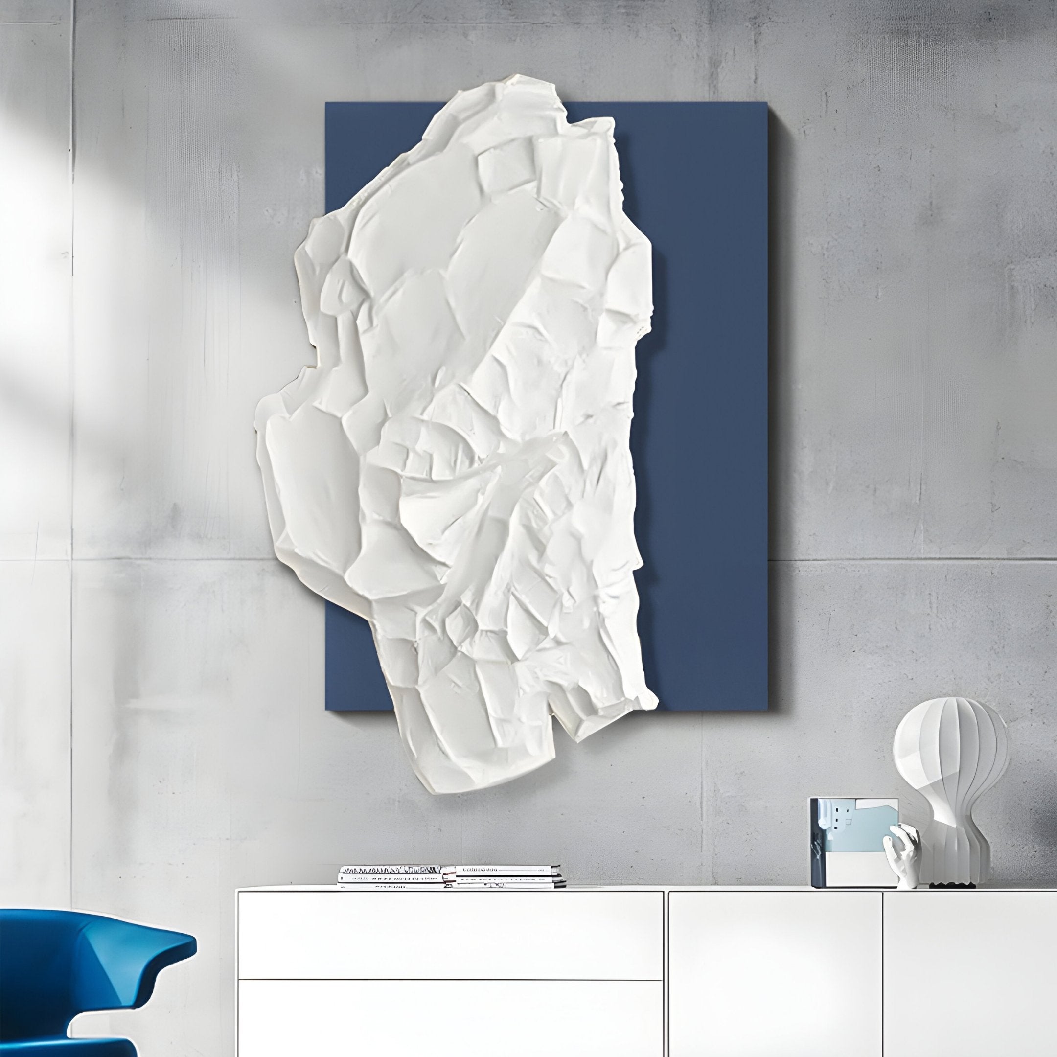 Displayed on a concrete wall is the Veritas White & Blue Abstract Wood Relief 3D Wall Art by Giant Sculptures, resembling crumpled paper. Below is a white cabinet with books and a modern lamp, with a blue chair partially visible in the corner.