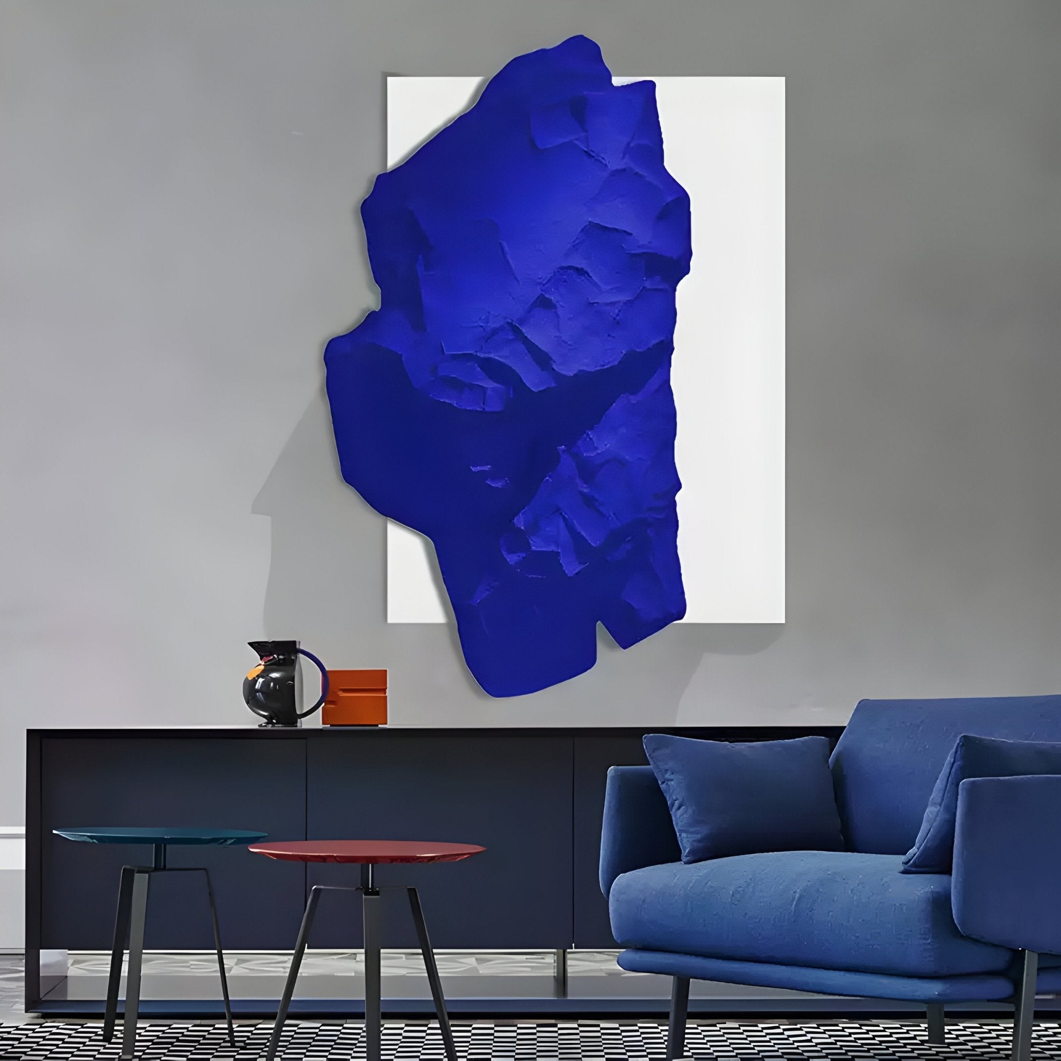 In the modern living room, a Veritas White & Sapphire Blue Wood Relief 3D Wall Art by Giant Sculptures adorns the gray wall. Below it sits a sleek black console table with a decorative object, while a blue couch and two small accent tables complete the contemporary setting.