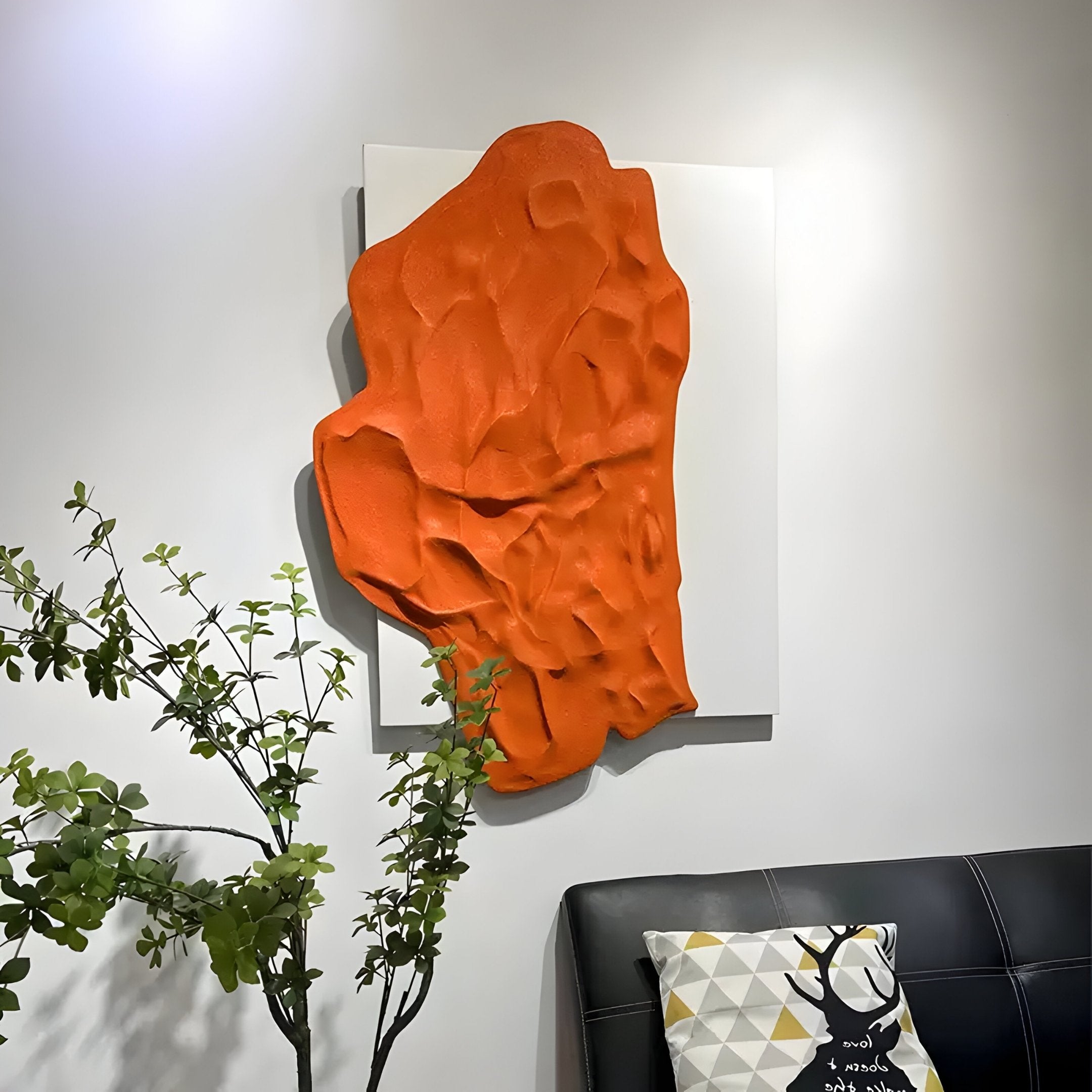 The Veritas White & Orange Wood Relief 3D Wall Art by Giant Sculptures adorns the wall above a black sofa with a geometric pillow, effortlessly integrating into the modern decor. A small green plant in the foreground offers a refreshing contrast to this vibrant scene.