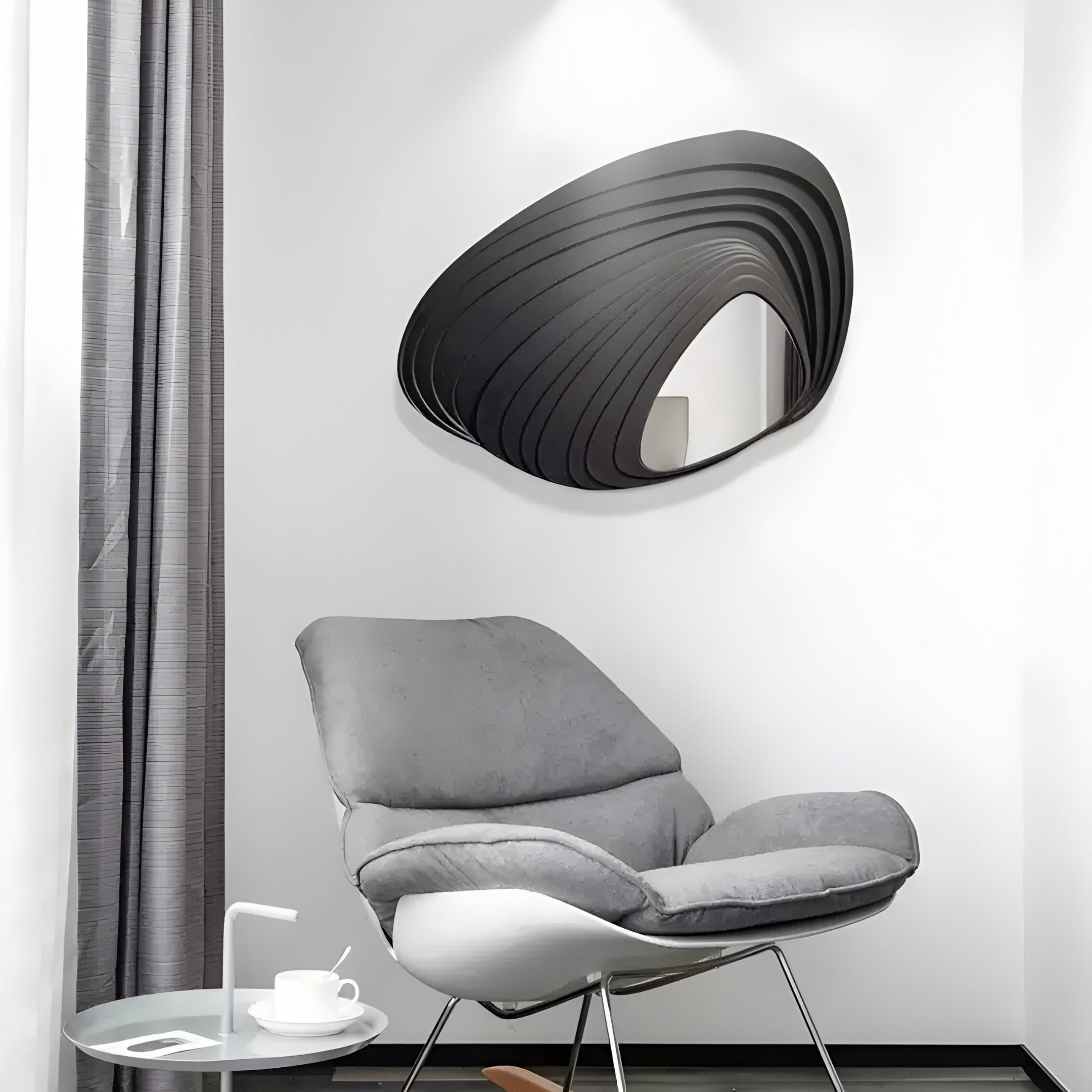 A modern interior showcases a sleek grey lounge chair, a small white side table with a cup, and the Reflecta Black Wood Carving Mirror 3D Wall Art by Giant Sculptures on the wall. A grey curtain adds softness to the minimalistic decor.