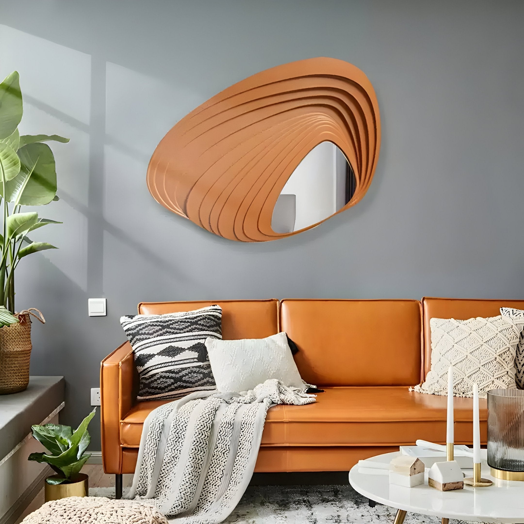 A modern living room showcases a sleek orange sofa with patterned pillows and a textured blanket. Above it hangs the Giant Sculptures Reflecta Orange Wood Carving Mirror 3D Wall Art. A potted plant and round coffee table with candles complete the inviting decor.