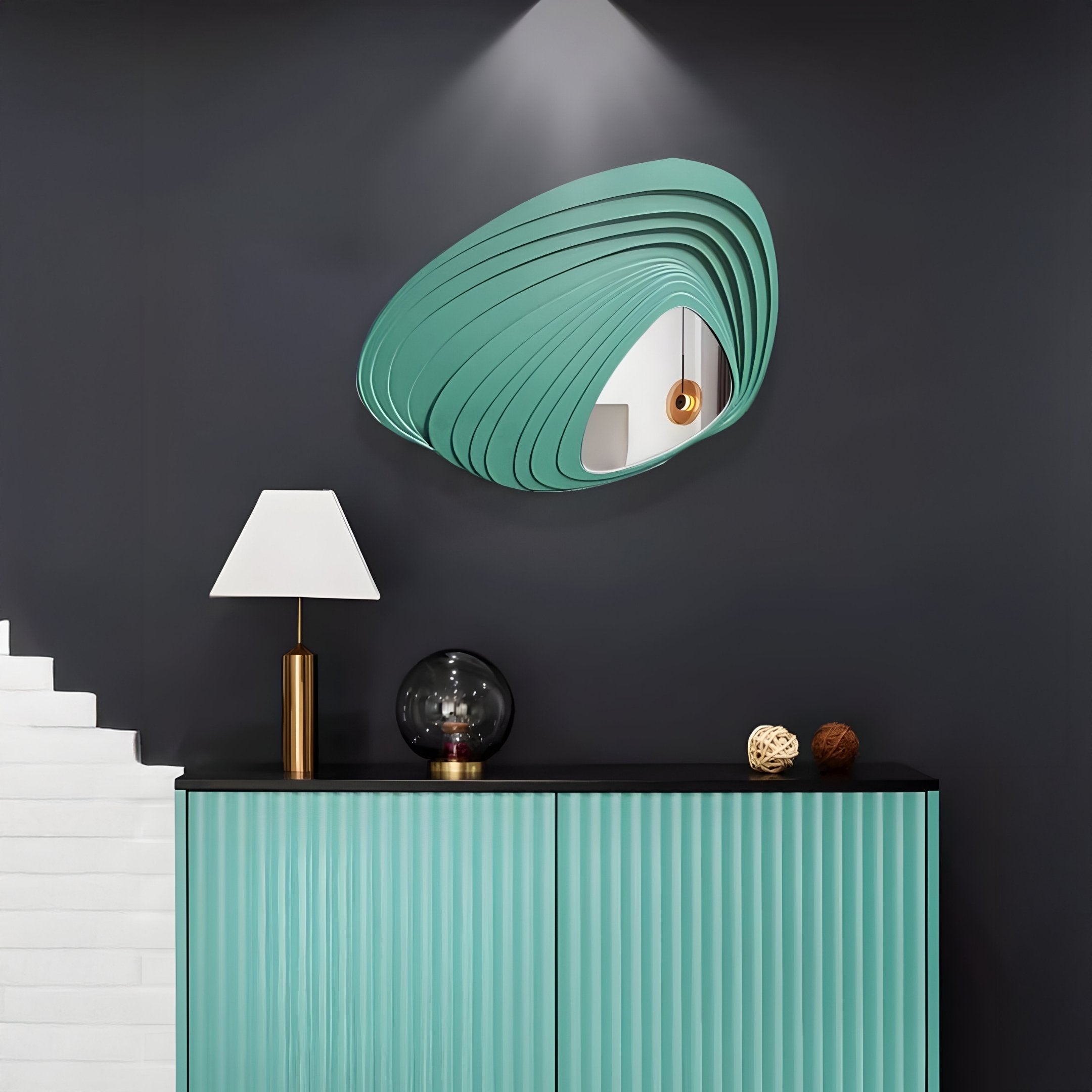 A modern interior features a teal cabinet against a dark wall, showcasing the Reflecta Green Wood Carving Mirror 3D Wall Art by Giant Sculptures. The cabinet holds a lamp with a white shade and decorative items, while partial white stairs on the left enhance the modern aesthetic.