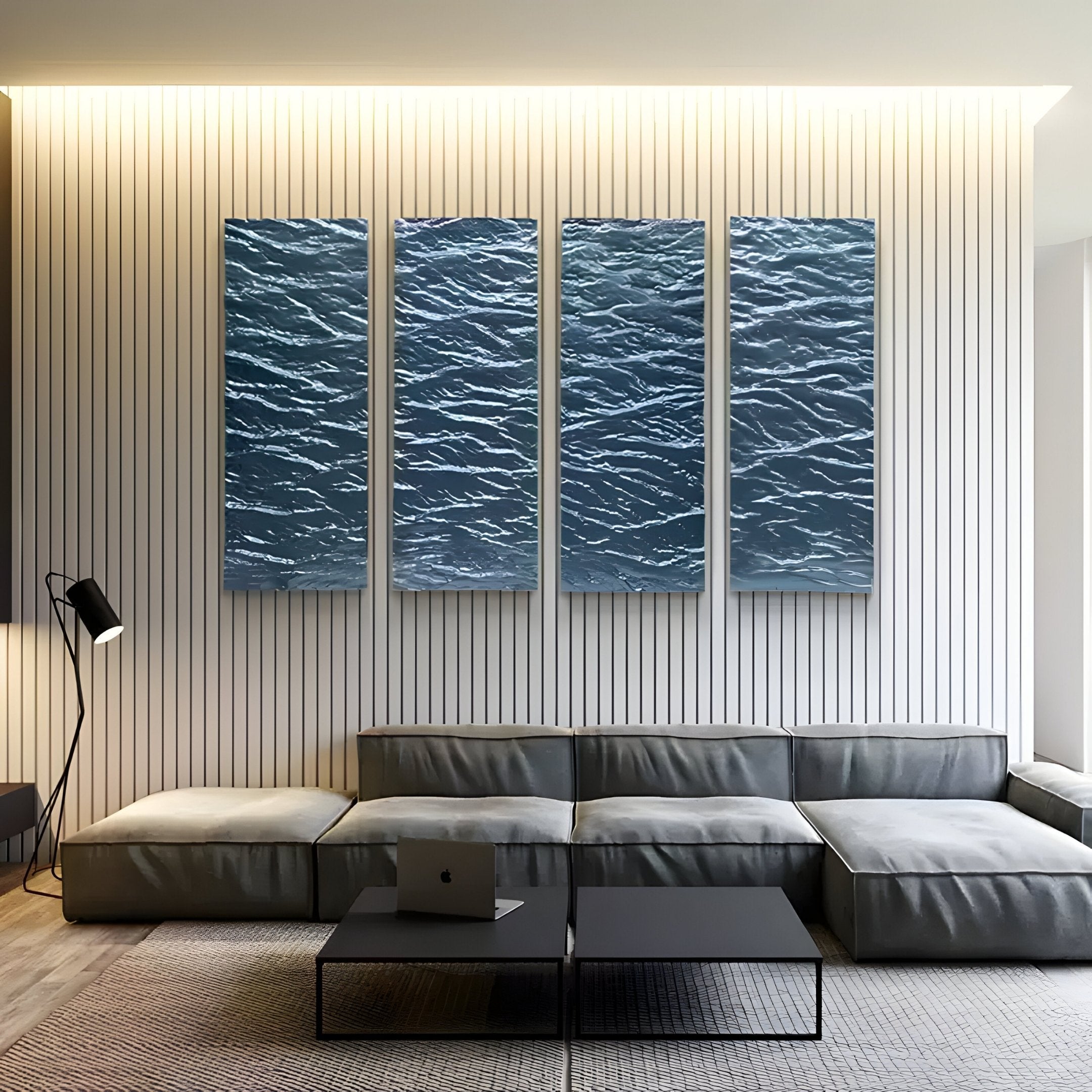 In a modern living room, a gray sectional sofa faces a wall with four vertical Illume 4-Panel Blue Hand-Painted Wood Relief 3D Wall Art pieces by Giant Sculptures, depicting rippling water. A floor lamp stands to the left; an open laptop sits on the black coffee table.