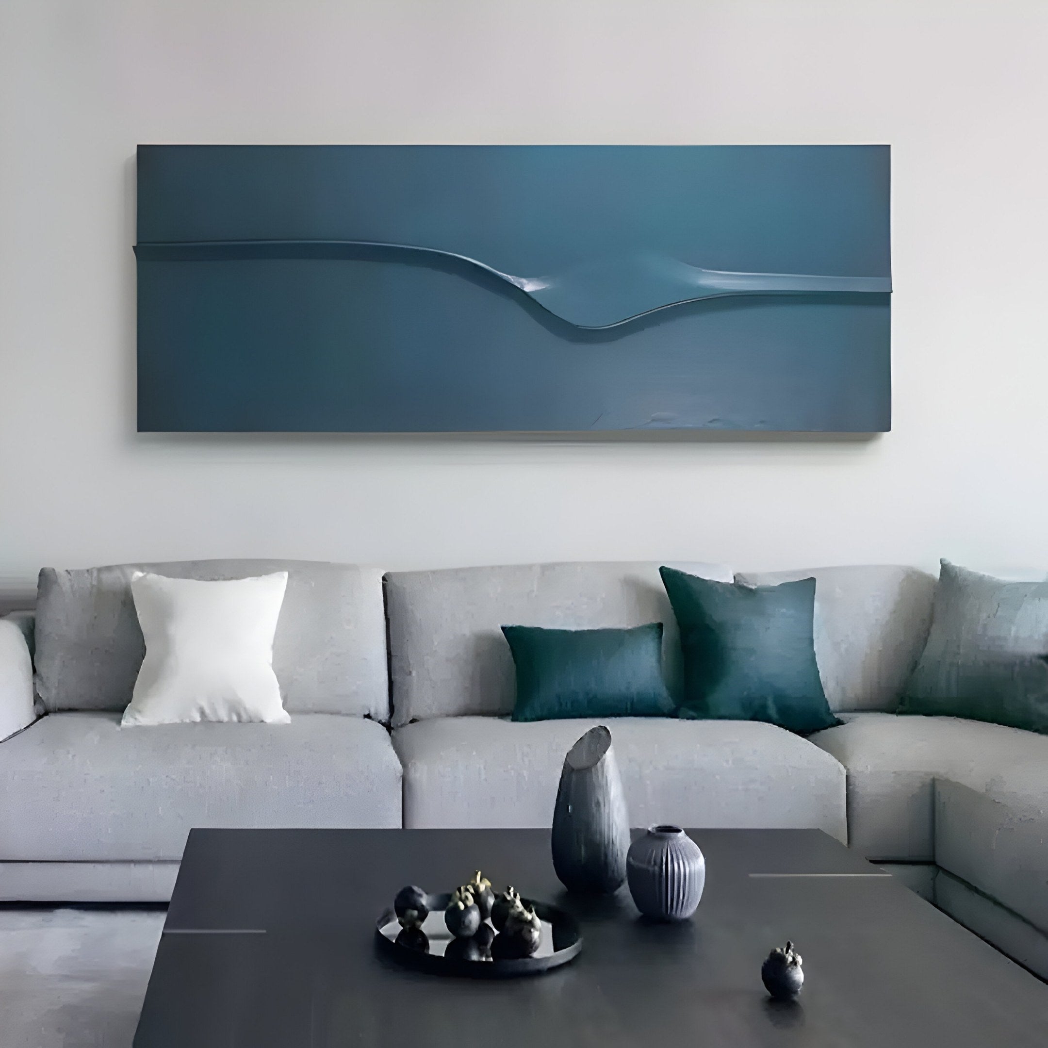 In a modern living room, a light gray sectional sofa with white and teal pillows is complemented by the Velar Horizontal Blue Wood Carving 3D Wall Art from Giant Sculptures showcasing a wave motif above it. A dark coffee table adorned with decorative vases and ornaments completes the scene.