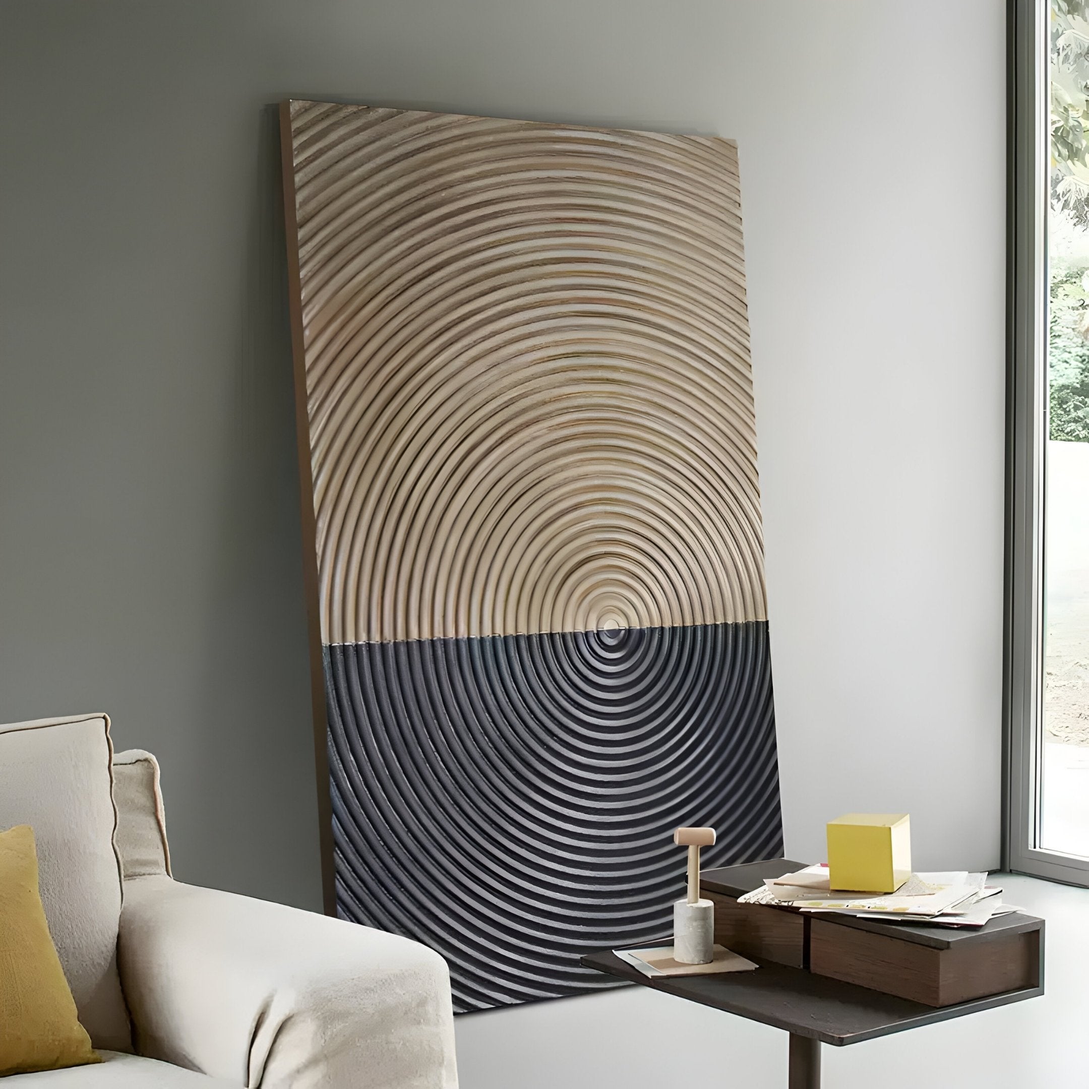 The Artura Beige & Black Hand-Painted 3D Relief Wall Art by Giant Sculptures, featuring concentric ridges, leans against a light gray wall near a window beside a beige sofa and a small table with books and a yellow box.