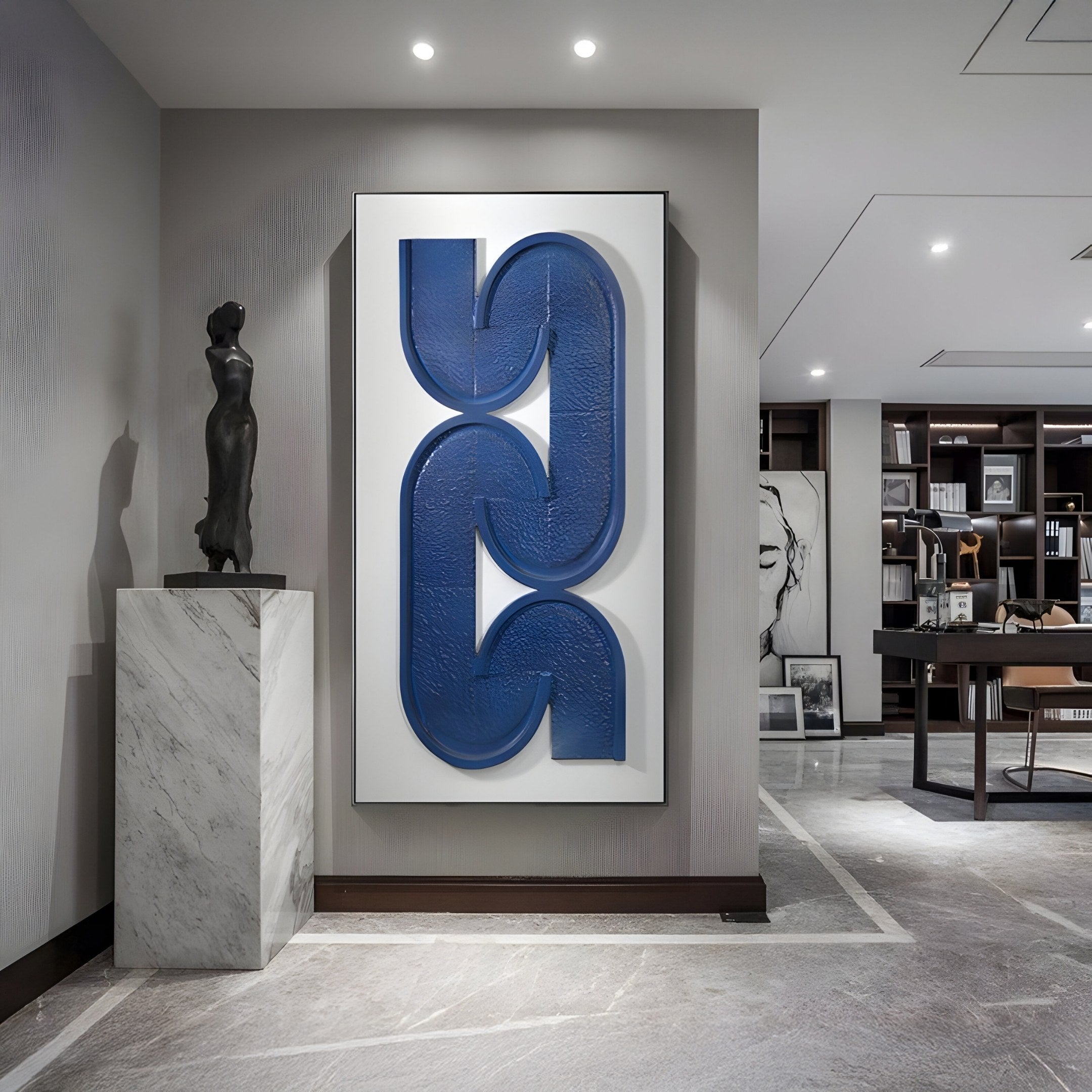 A modern interior features the Infinique Vertical Blue Abstract Wood Carving 3D Wall Art. To the left, a marble pedestal holds a Giant Sculptures dark piece, contrasting against wooden shelves in the workspace background adorned with framed art.