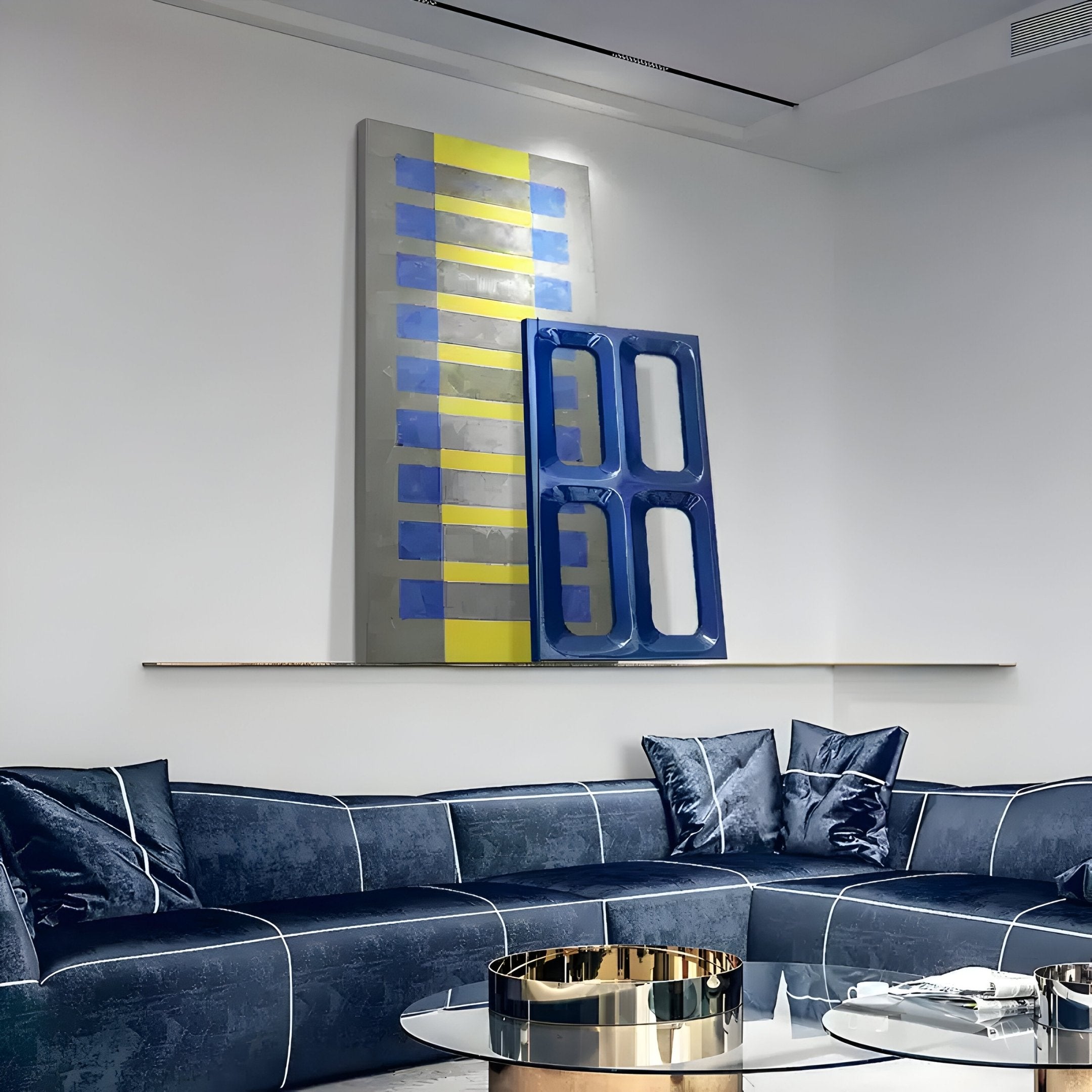 In a contemporary living room, a dark blue sectional sofa with decorative pillows is complemented by Giant Sculptures Lineris Blue Oil Painting & Glossy Wood Relief 3D Wall Art in blue and yellow stripes. A round glass coffee table with a gold tray stands prominently in the foreground.