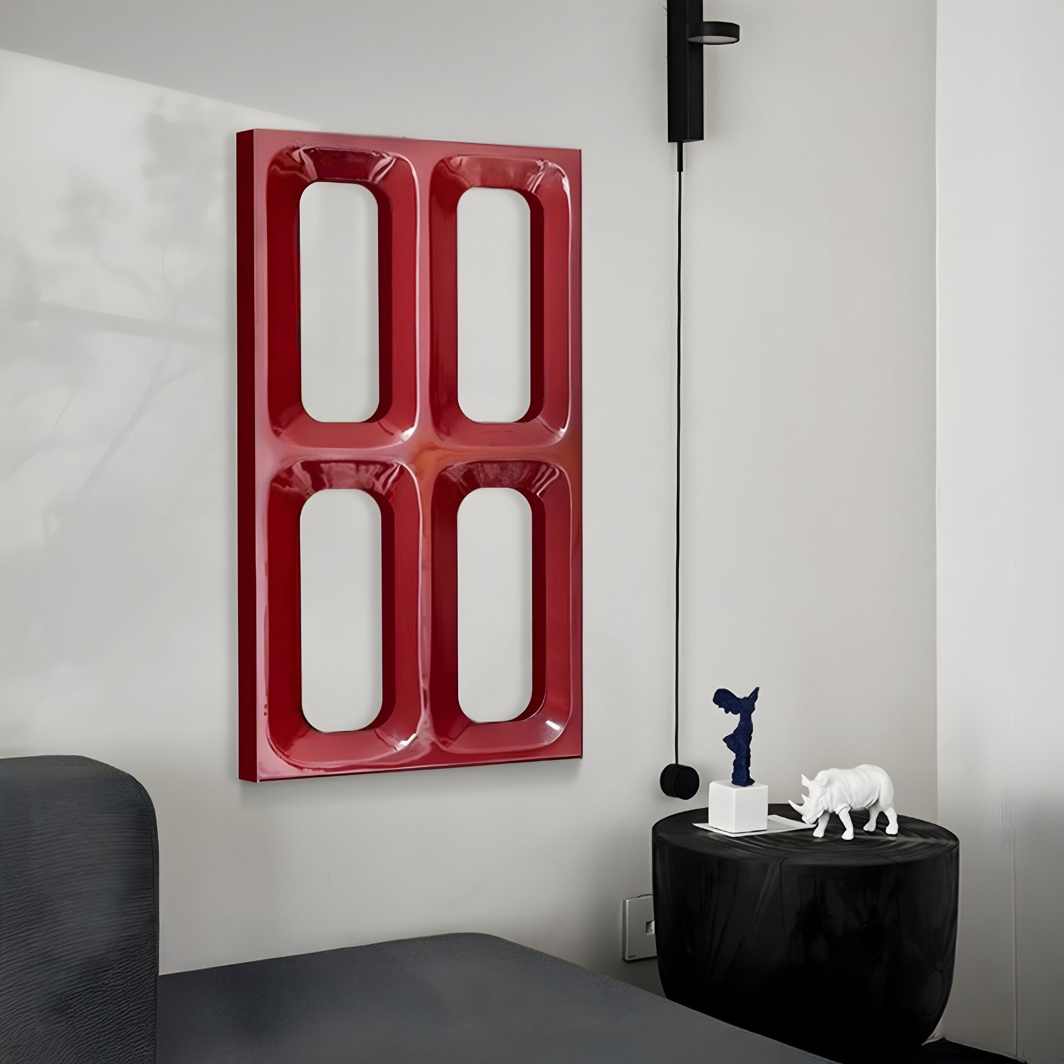 A modern room showcases the Lineris Red Glossy Wood Relief 3D Wall Art by Giant Sculptures with large, rounded cutouts. Below, a small black side table holds two abstract sculptures: a blue figure and a white rhinoceros. To the left is a dark sofa completing the sleek aesthetic of modern interiors.