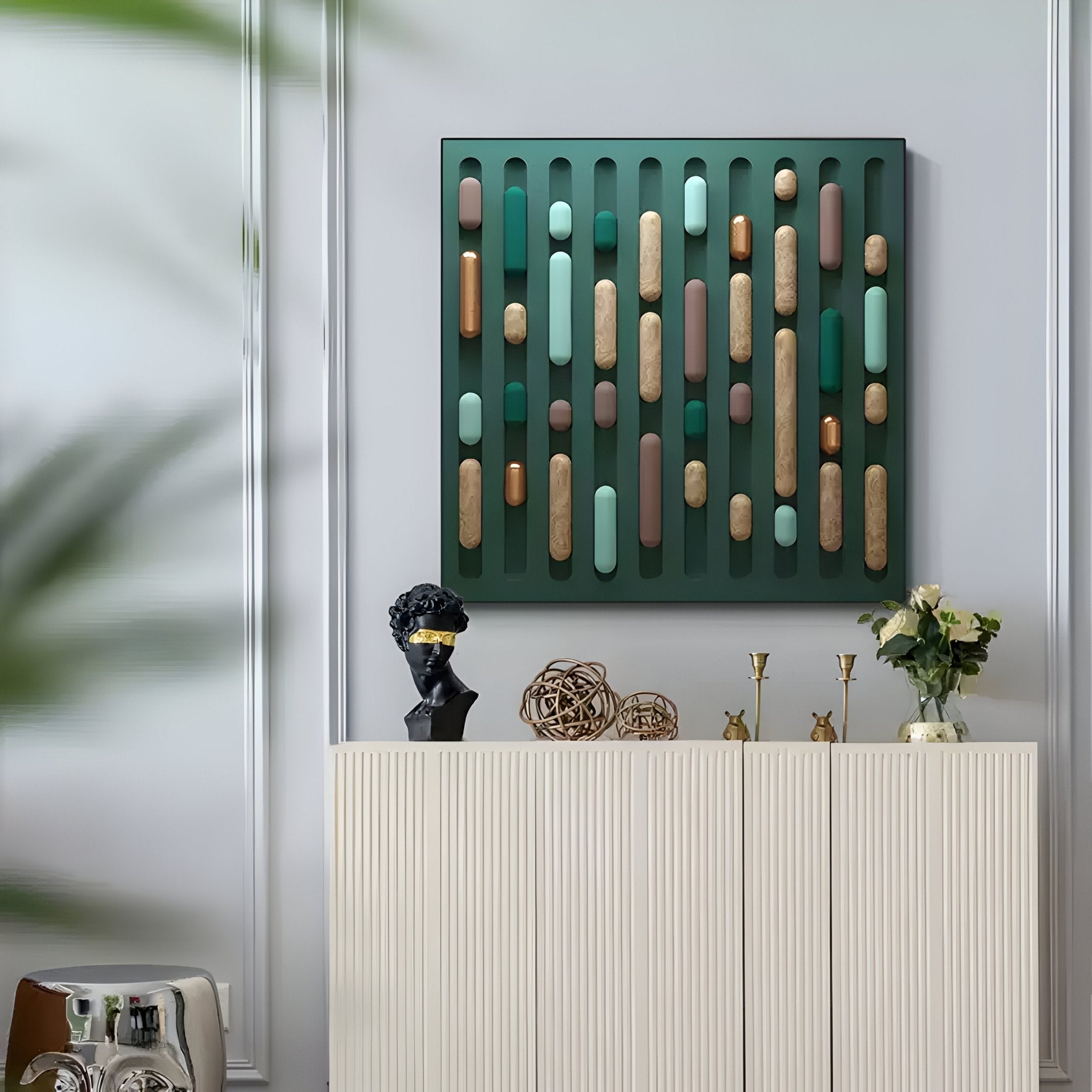 In a modern, Nordic-inspired living room, a white cabinet adorned with gold decor and a black bust stands beneath Giant Sculptures Formexa Capsule Geometric Wood Relief 3D Wall Art. This green abstract piece of cylindrical shapes hangs on the gray wall, while a blurred plant leaf appears in the foreground.