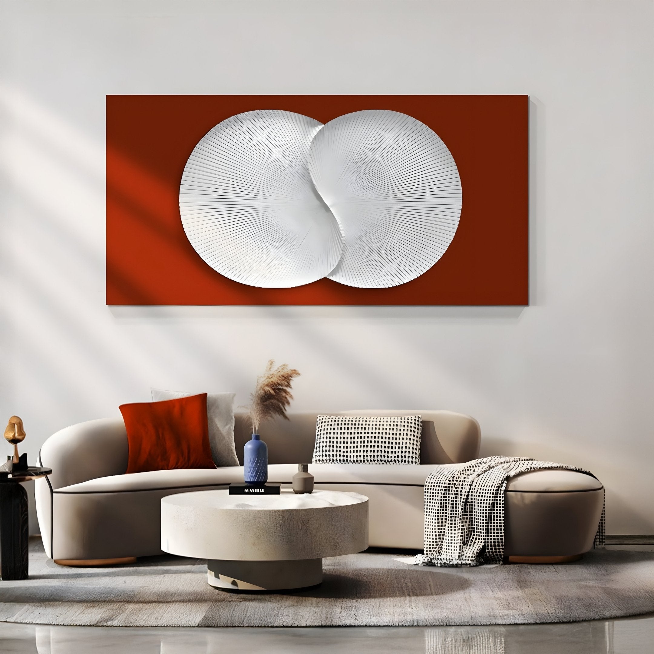 A modern living room showcases a curved beige sofa with orange and patterned cushions. A round white coffee table anchors the space, while the Artivio Red & White Wood Relief 3D Wall Art by Giant Sculptures adds flair above. Sunlit shadows and pampas grass enhance the decor.