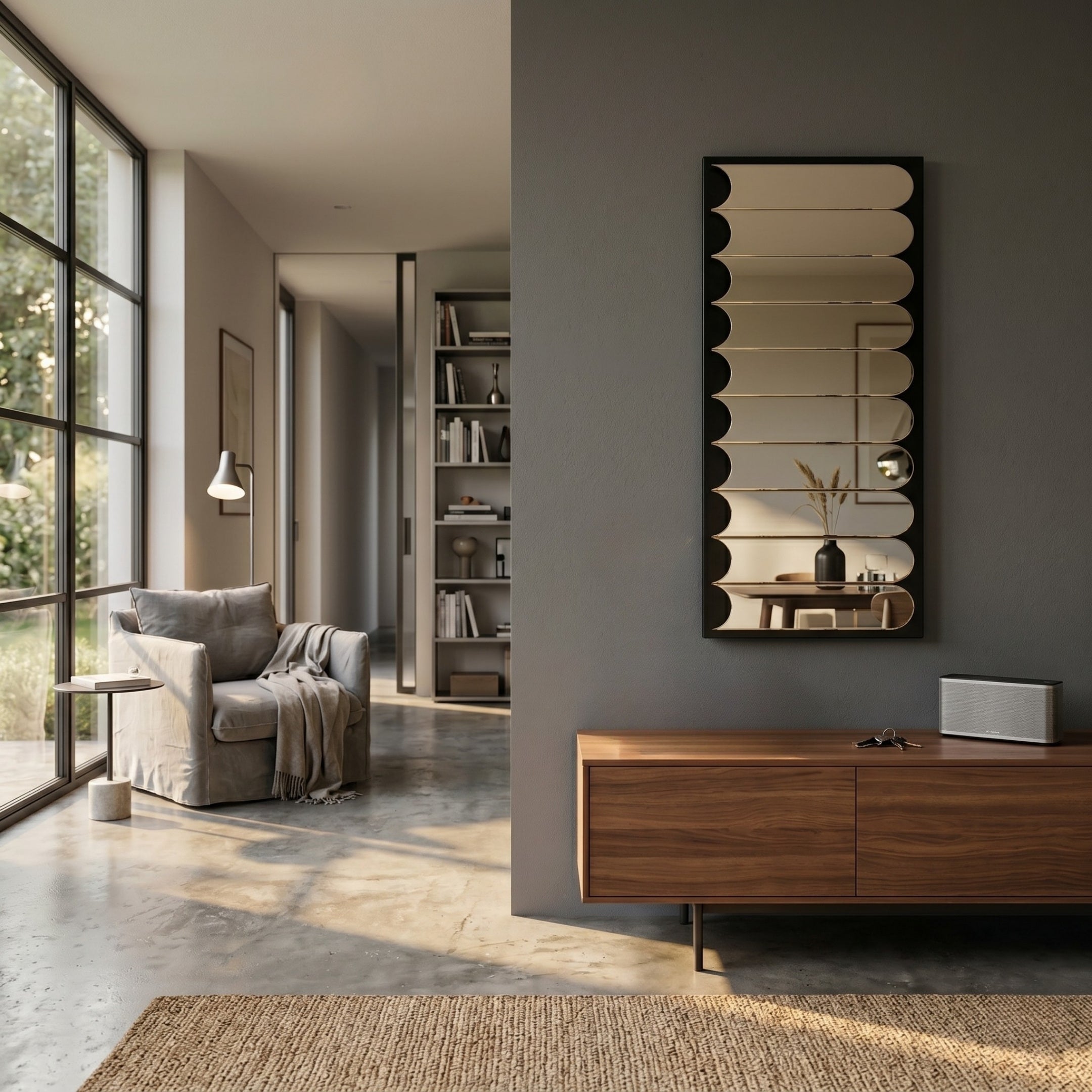 A modern living room with large windows, a gray armchair draped with a blanket, a wooden sideboard, and the Giant Sculptures Lumiscale Black & Taupe Modular Glass Wall Mirror on a dark wall. Sunlight fills the space with warmth.