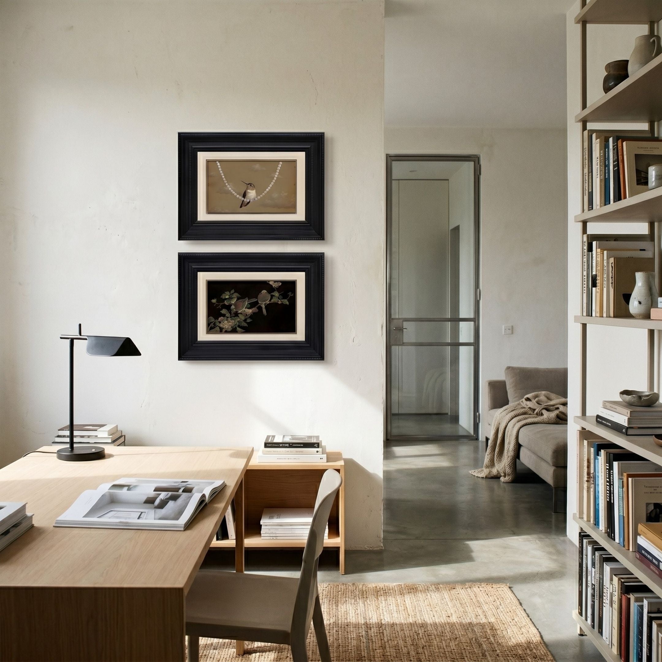 A modern home office features a wooden desk, chair, black desk lamp, and books. Two Kansora Songbird Pearl & Branch 2-Set Framed Wall Art pieces by Giant Sculptures hang on a white wall near shelves with books and decor, overlooking a cozy living area.