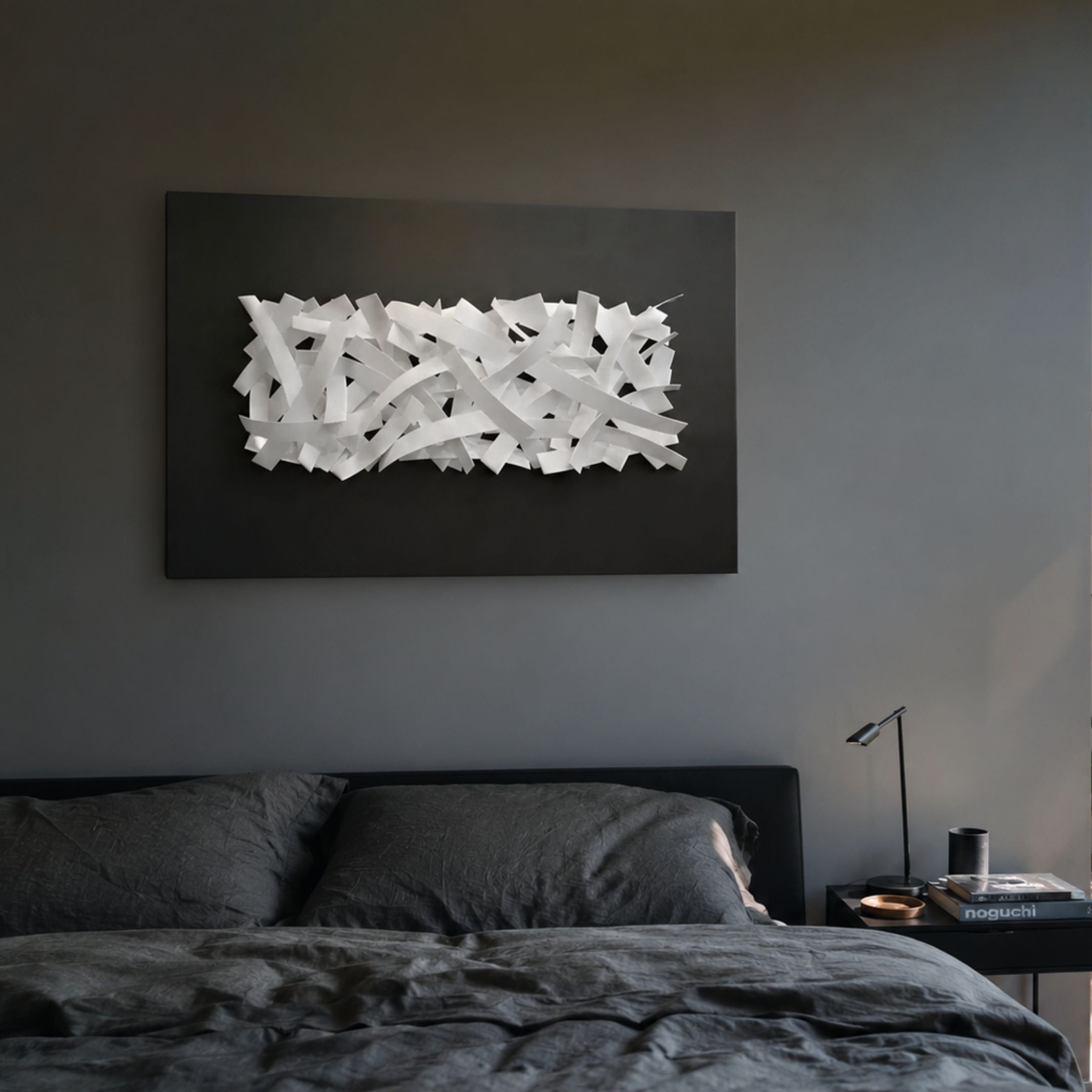 Stravira Black I Sculptural White Metal 3D Wall Art with interwoven white iron strips on a black panel, mounted above a dark grey bed and nightstand.