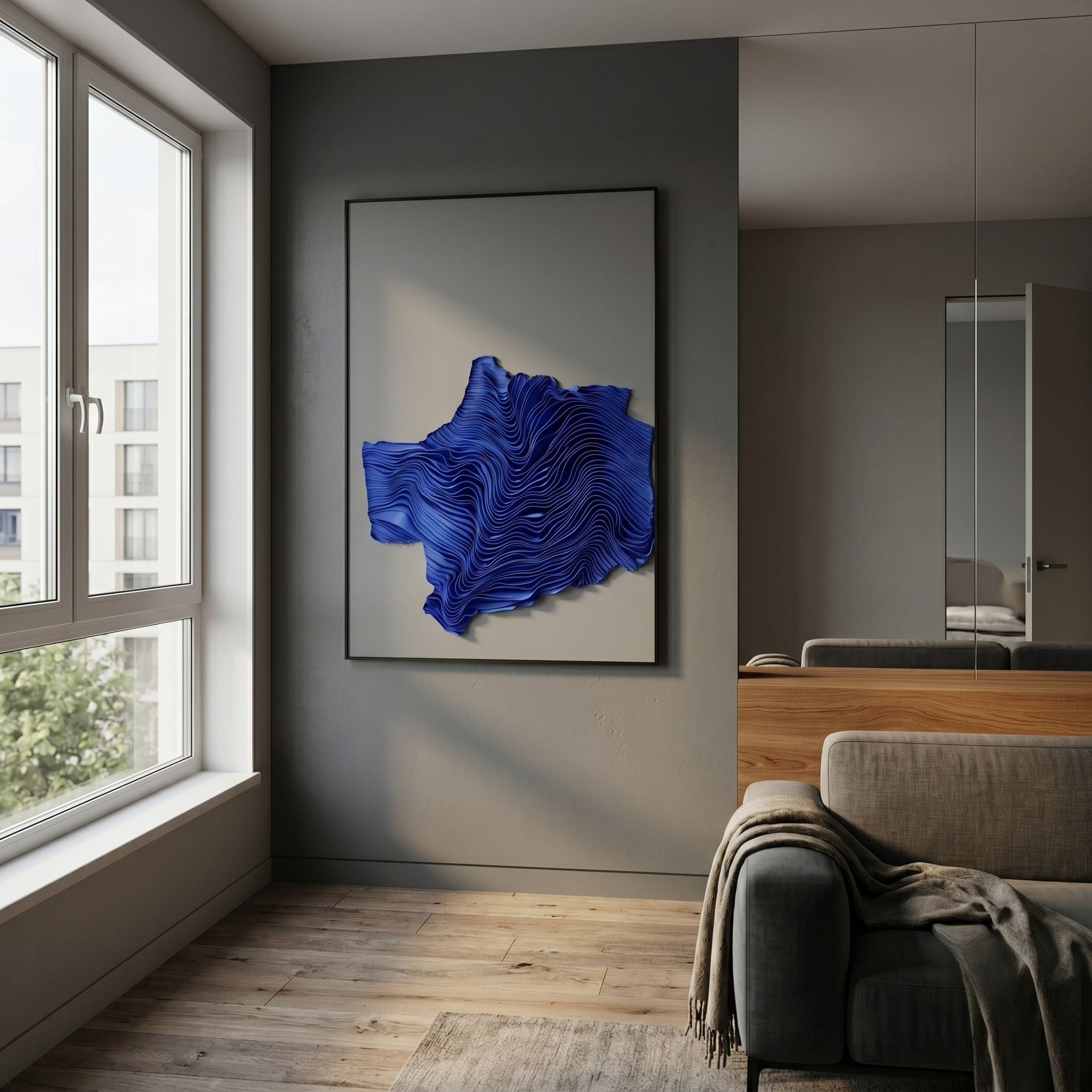 Ravelle Blue Crest Felt 3D Relief Wall Art in a black frame, displayed on a grey wall in a modern living room beside a window and grey sofa.