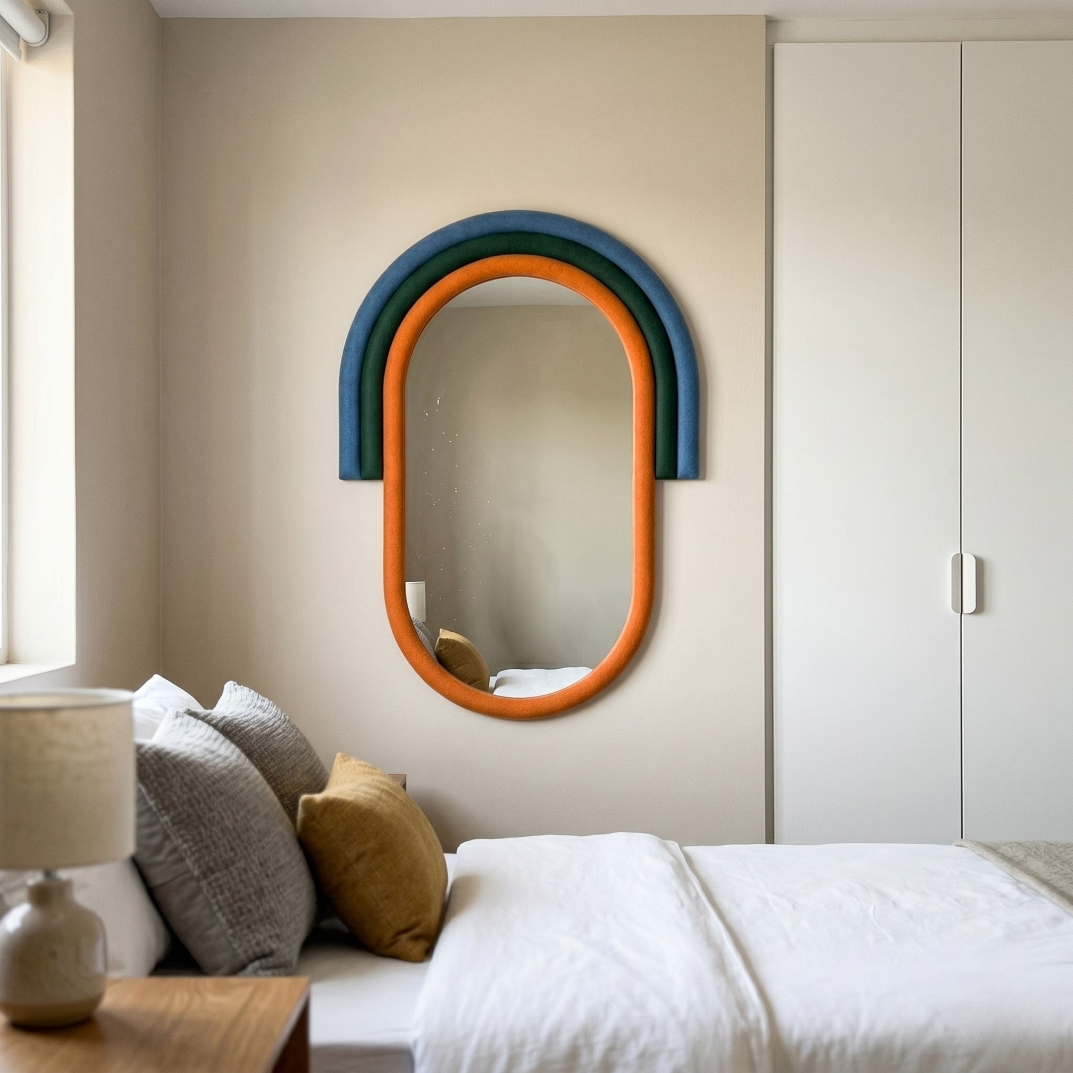 A modern bedroom features a neatly made bed with white bedding, gray and mustard pillows, a bedside table with a lamp, and the Arcaluna Blue & Orange Layered Arch Mirror 3D Wall Art by Giant Sculptures on a beige wall.