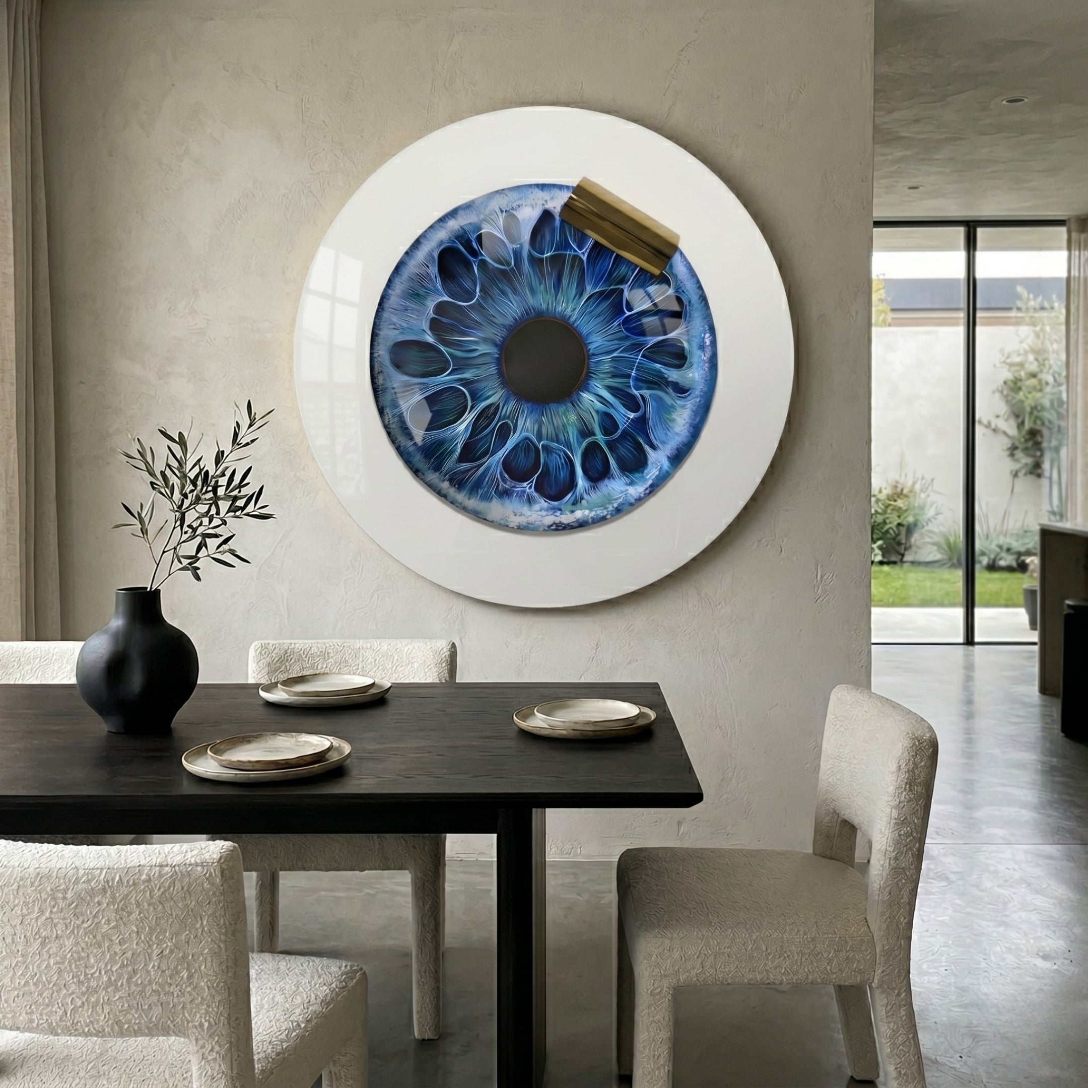 A modern dining room features a dark table, white chairs, and the Iriselle Oceanic Halo Crystal Porcelain Round 3D Wall Art by Giant Sculptures, resembling a blue eye. A black vase with greenery sits on the table as sunlight filters through sheer curtains.