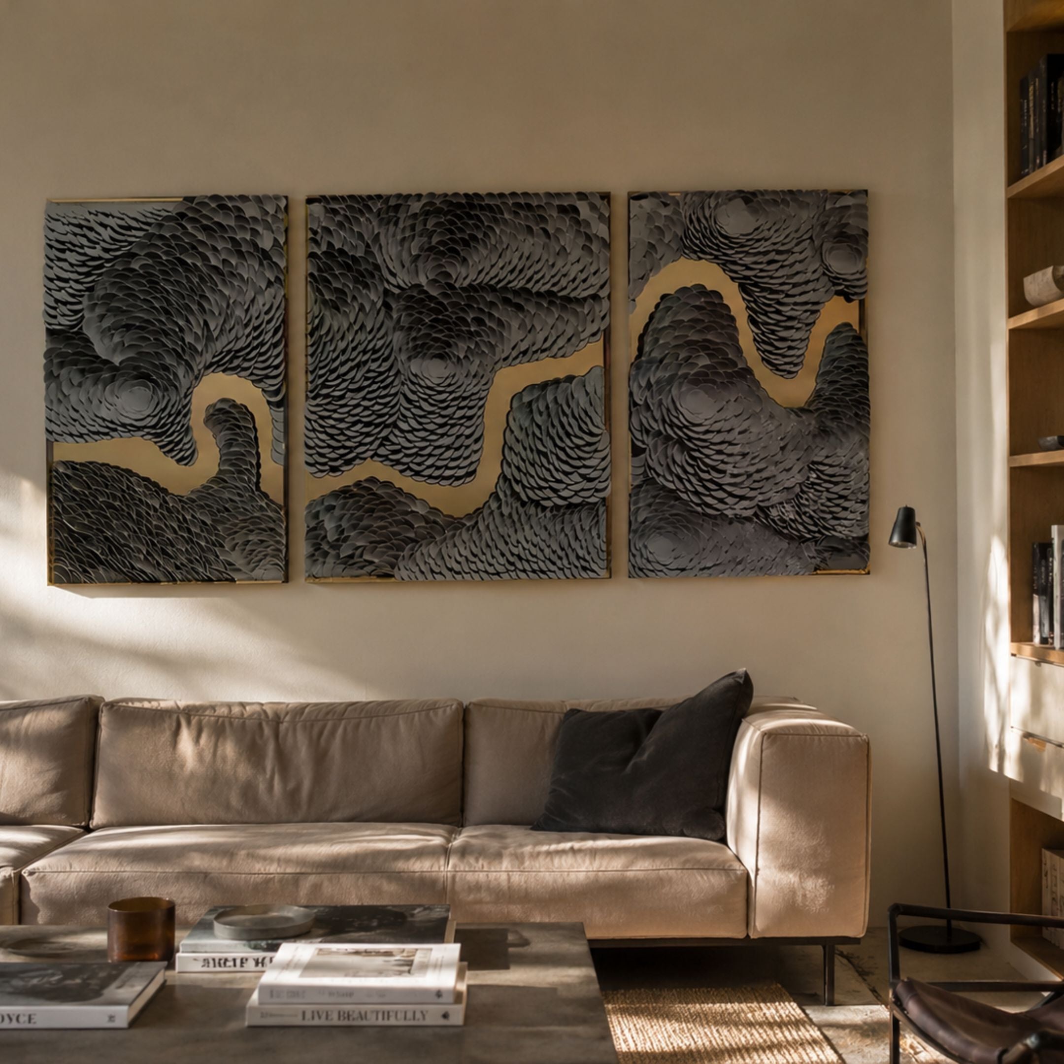 Riverine Flow Charcoal Grey Textured Triptych Wall Art by Giant Sculptures hung above a beige leather sofa in a sunlit living room with bookshelves.