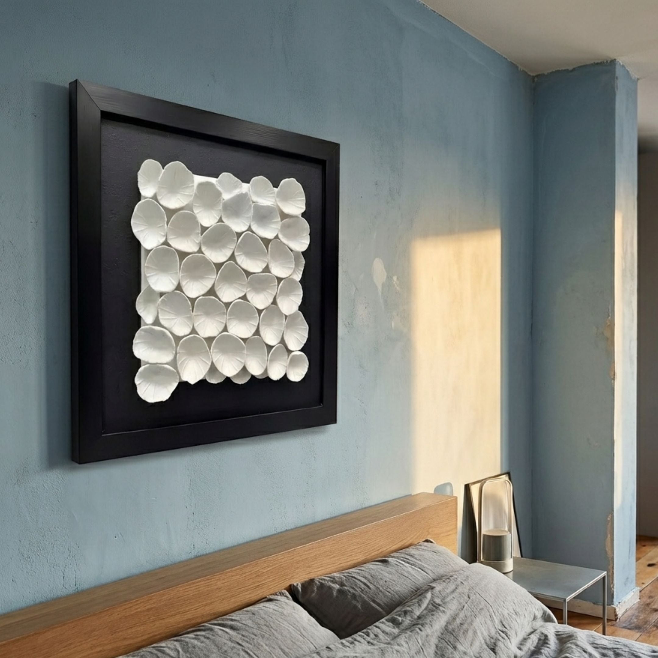 Harmonia 3D Ceramic Petal Wall Sculpture with black frame mounted on a blue bedroom wall above a wooden headboard with grey bedding and side table.