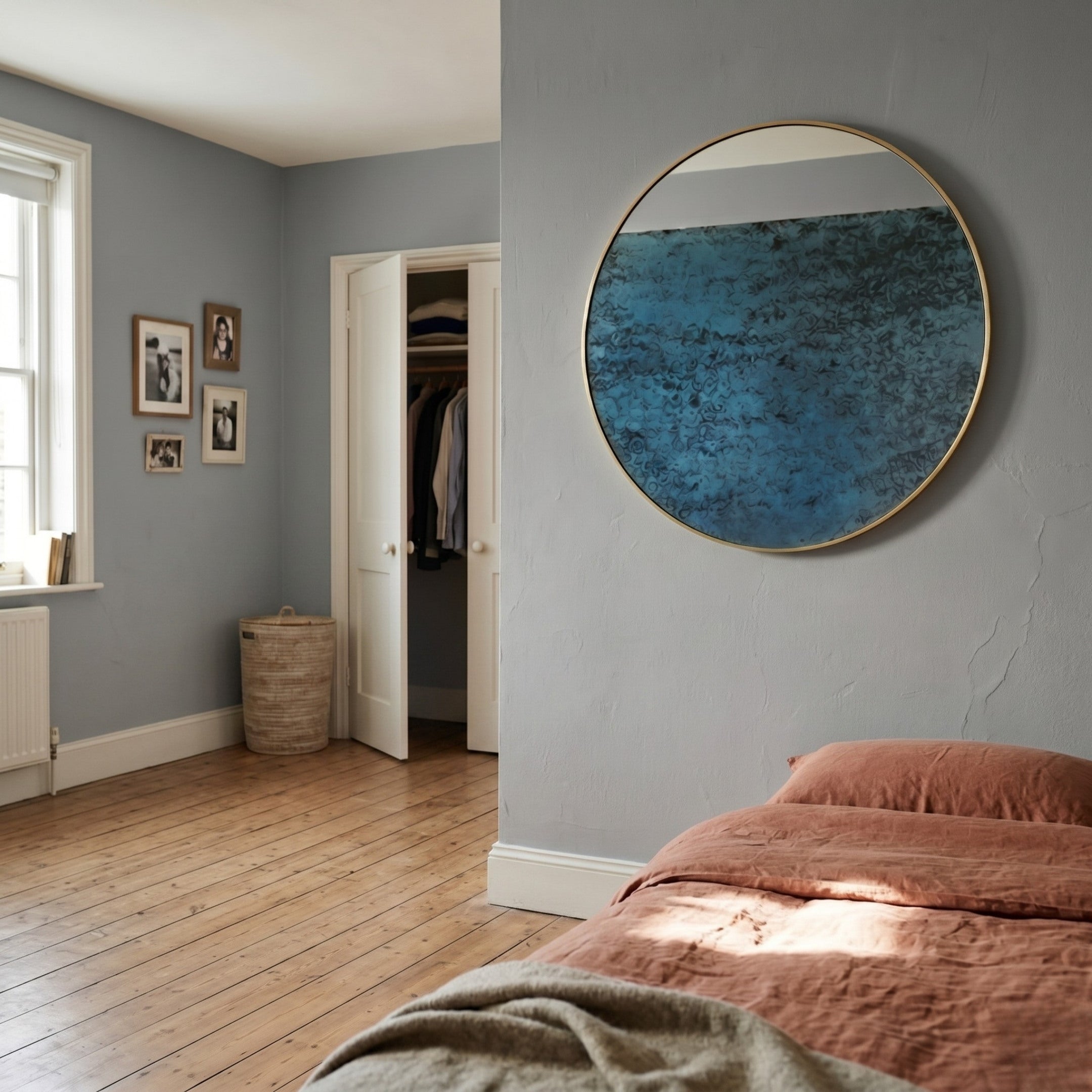 A cozy bedroom with light blue walls, a round mirror, wooden floor, rust bedding, framed photos, and the Auralune Blue Horizon Abstract Round 3D Wall Art by Giant Sculptures above the bed; plus a closet door and woven laundry basket by a sunlit window.