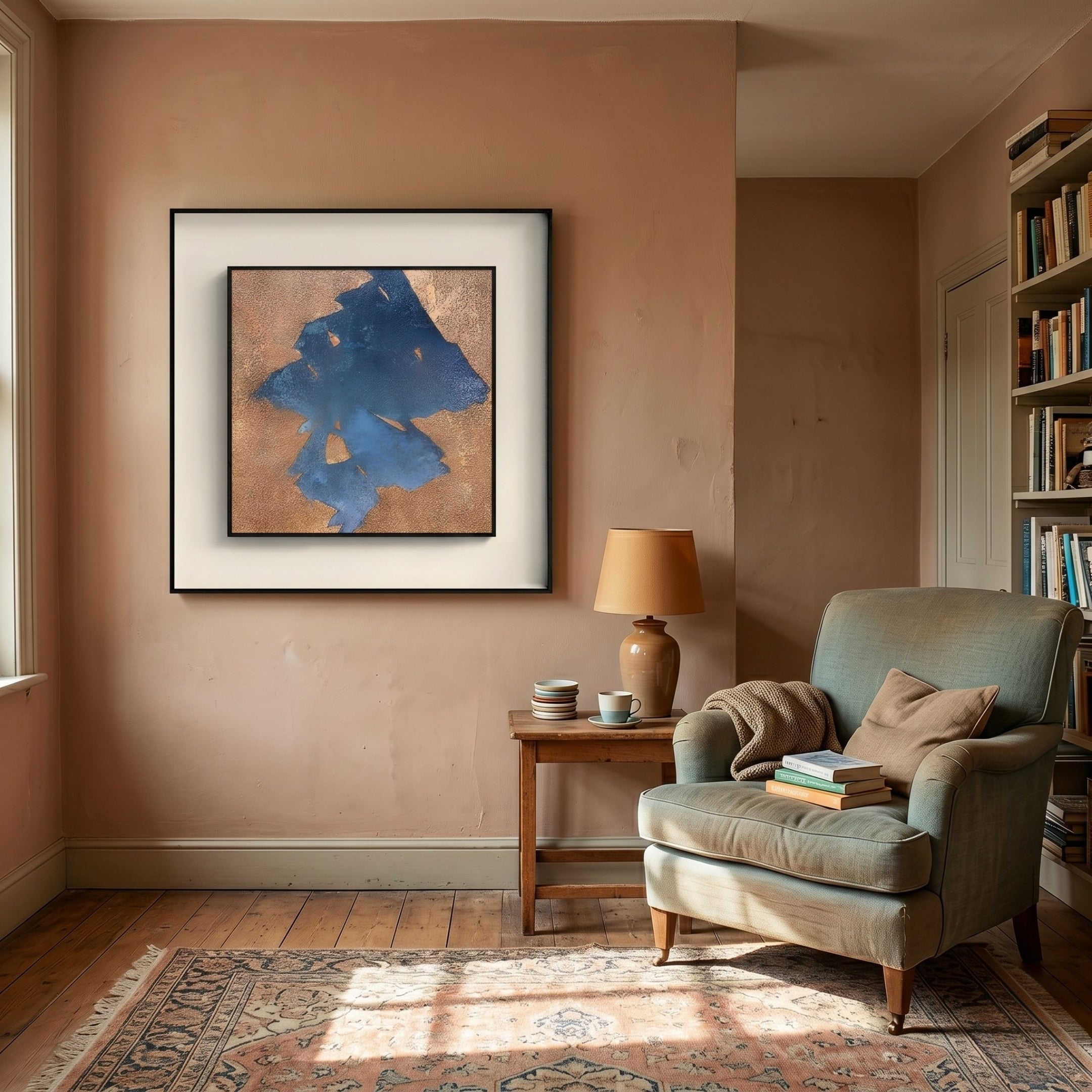 A cozy reading nook features an armchair, books, and a lamp on a side table. Above hangs the Stratum Sandform Blue Textured Abstract Framed Wall Art by Giant Sculptures. A bookshelf is on the right, and a patterned rug covers the wooden floor.