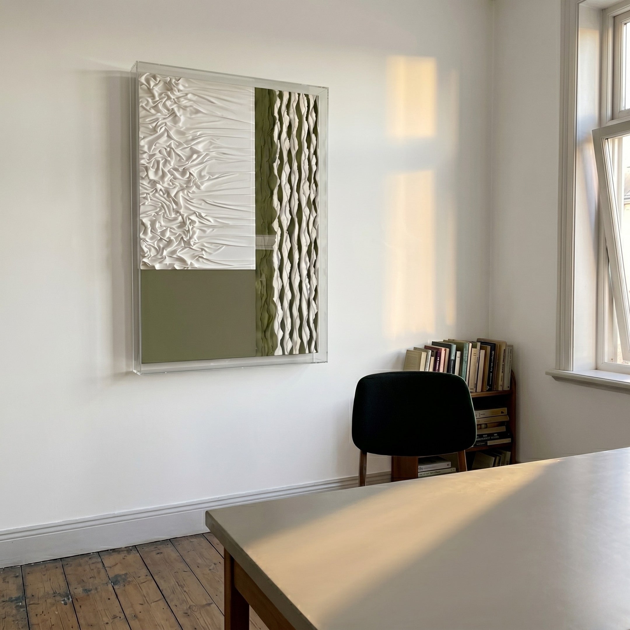 A minimalist room with wooden floors, a table, and a black chair by a small bookshelf under a window features Giant Sculptures’ Texturien White & Olive Paper Abstract 3D Wall Art on the wall, illuminated by soft sunlight.