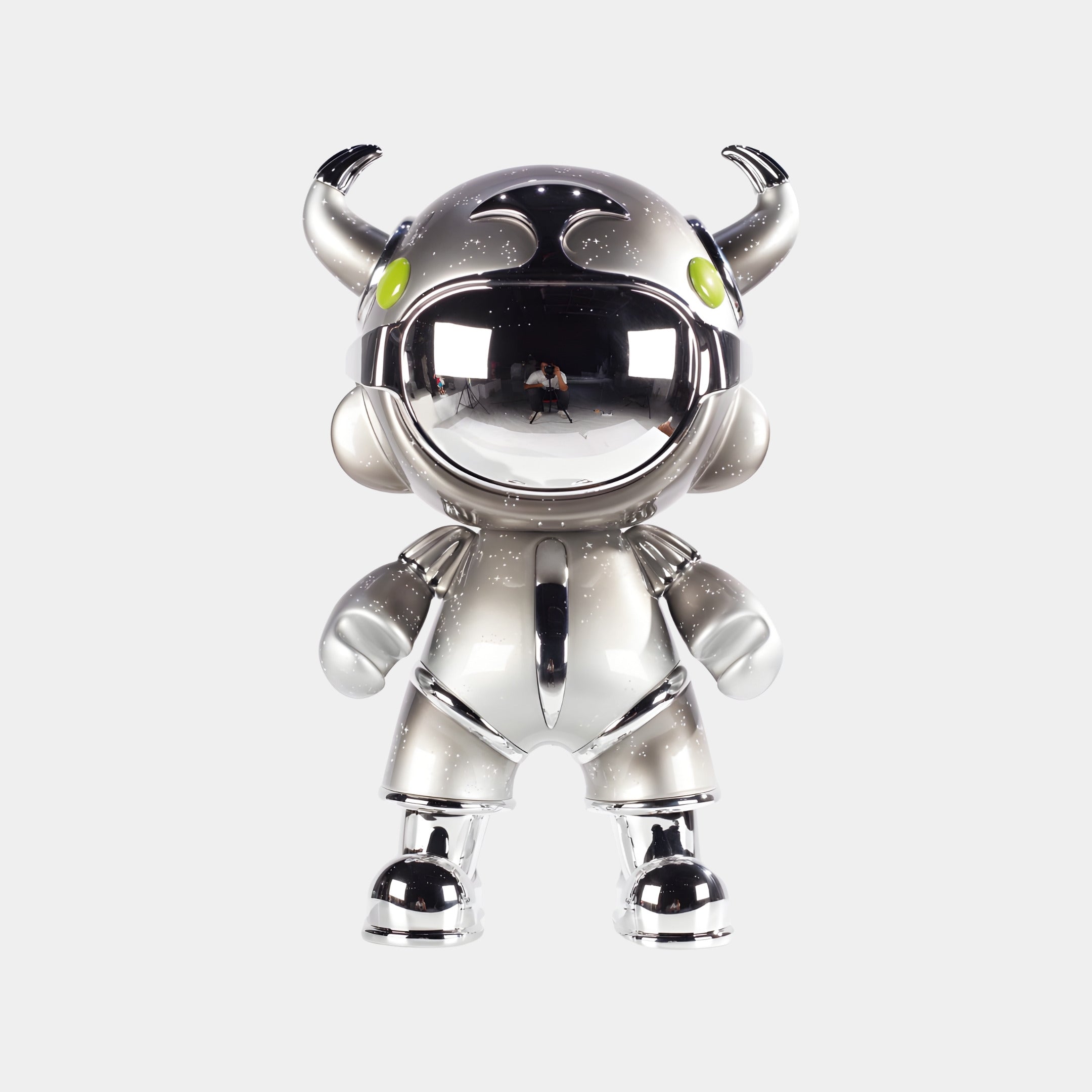 The Giant Sculptures Platinum Suit Astro Devil Sculpture - 80cm is a shiny, metallic humanoid with a large round head, horns, bright green eyes, short limbs, mirrored chrome visor, and a striking metallic gradient finish on white.