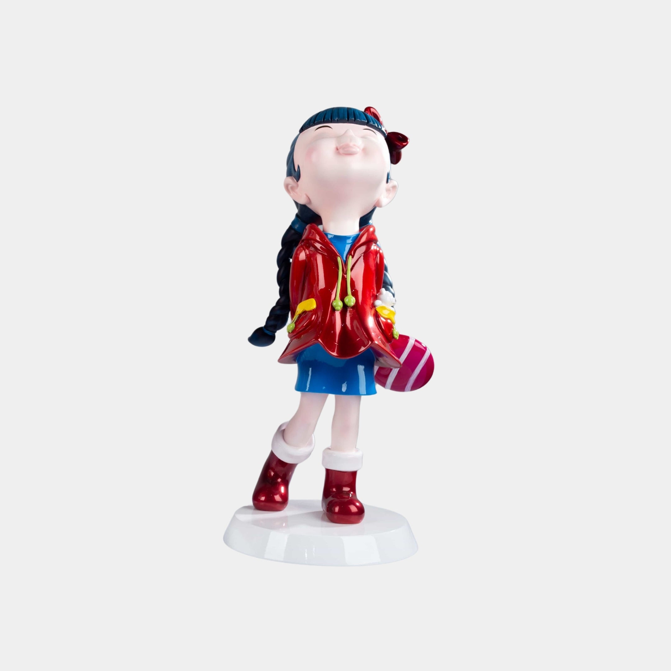 The Cherry Red Lulabelle Sculpture - 60cm by Giant Sculptures features a smiling girl with blue braided hair, red jacket and boots, blue skirt, and a pink-striped gift box—a vibrant decorative statement on a white base.