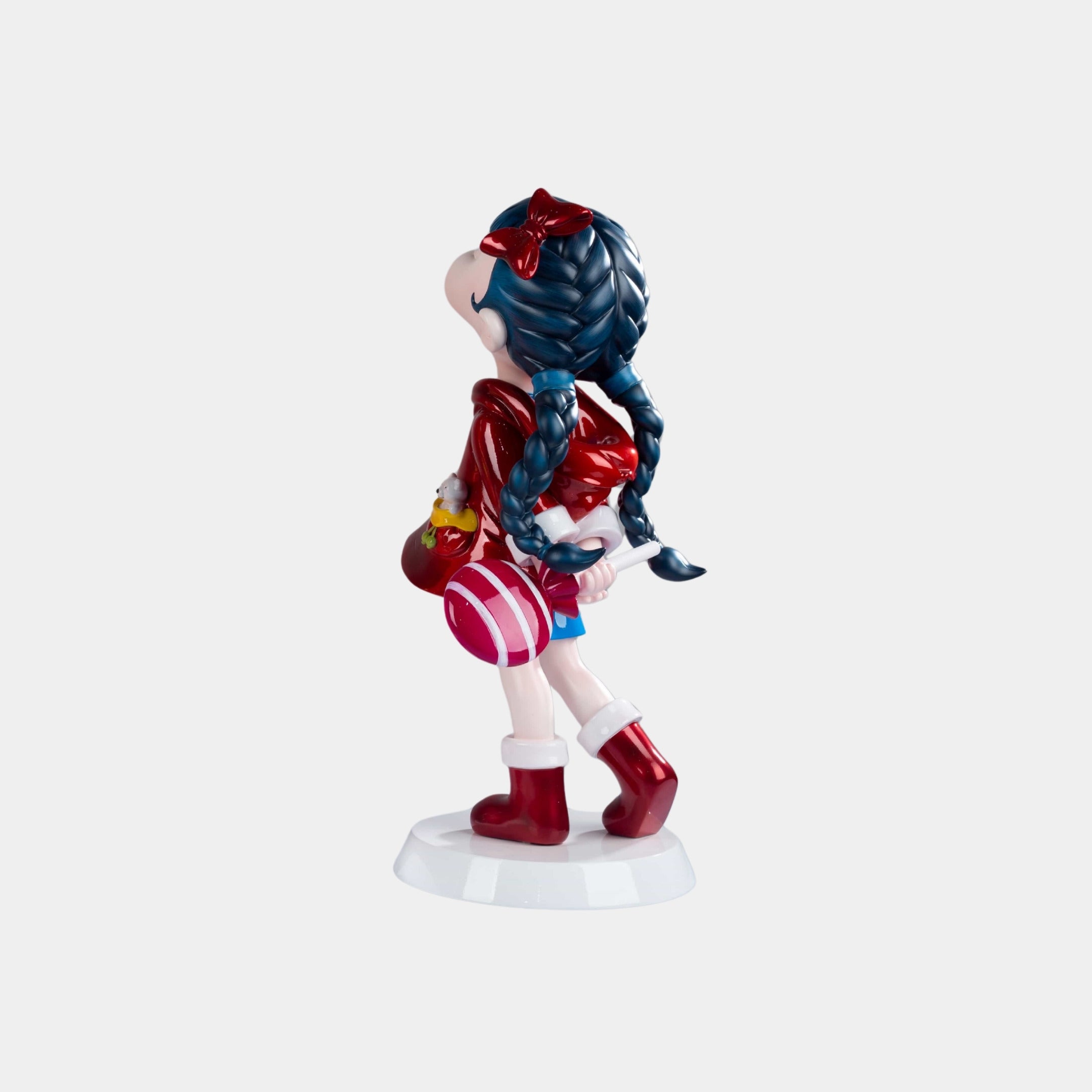 The Cherry Red Lulabelle Sculpture - 60cm by Giant Sculptures features a girl with blue braided hair, red bow, jacket, boots, blue skirt, and pink-striped ball—an expressive side-view statement piece on a white base.