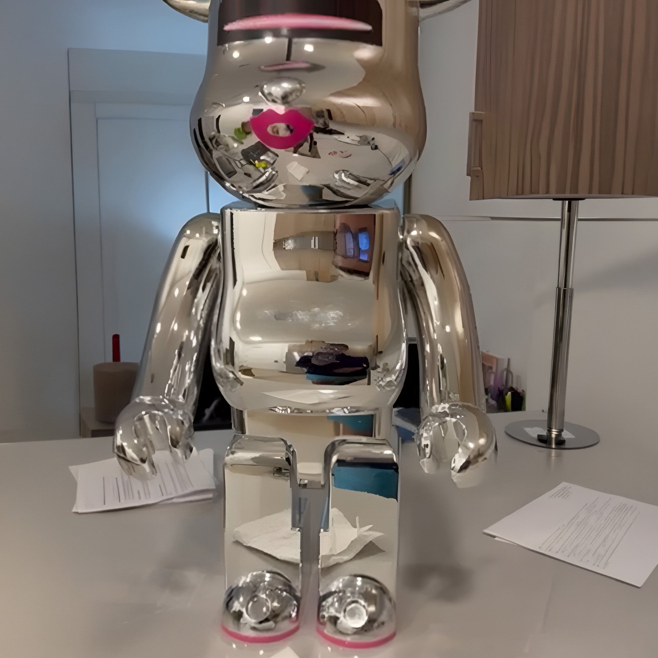 The Neon Kiss Chrome Bear Sculpture - 70cm (Ex Display) by Giant Sculptures, with its mirror finish and pink details, adds playful pop-art flair to a modern office desk, reflecting surroundings among papers and a lamp.