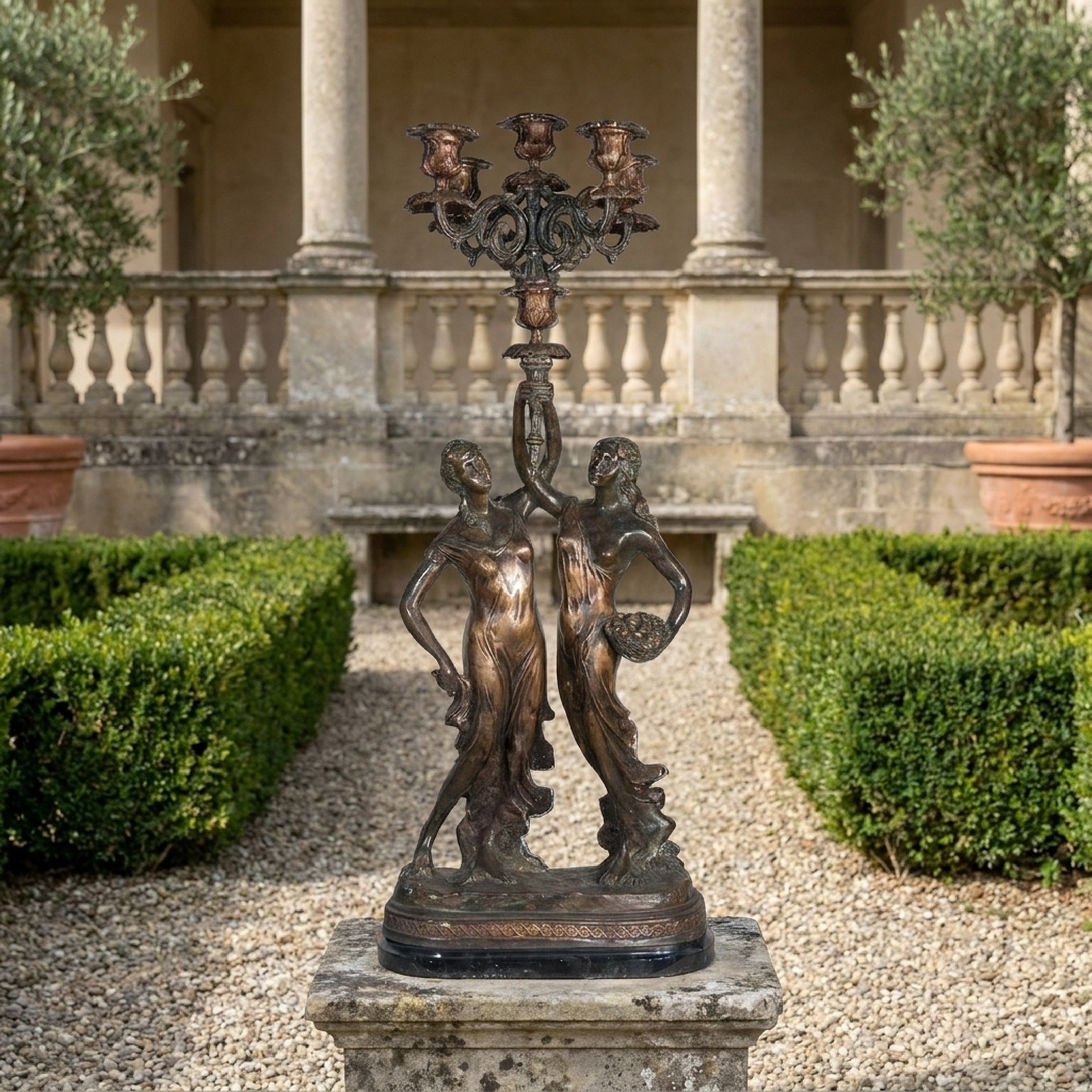 Giant Sculptures' Classical Two Female Figures Candelabra Bronze Sculpture on a stone plinth in a formal garden with hedges and a balustraded villa behind.