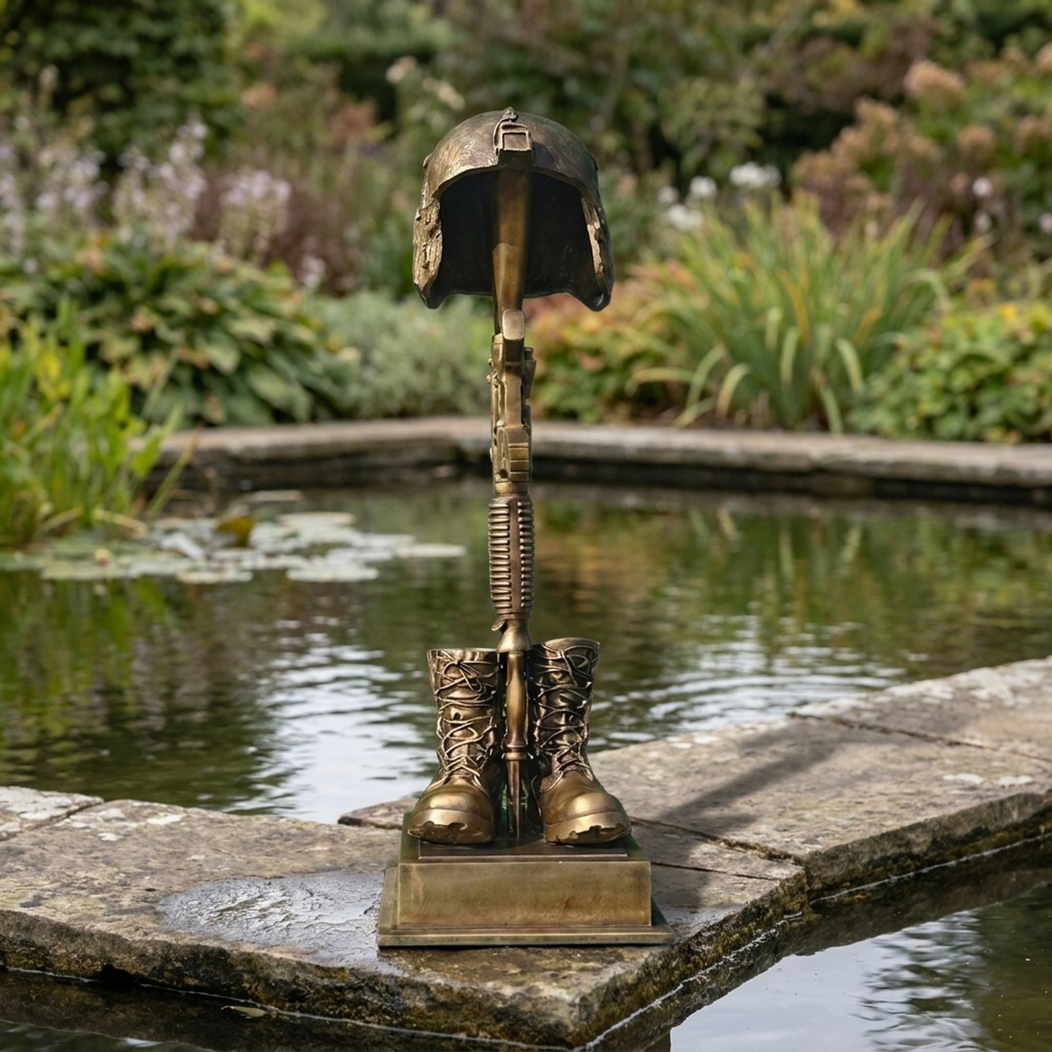 Contemporary Battle Cross Memorial Bronze Sculpture by Giant Sculptures, with helmet, rifle and boots on a stepped plinth, displayed beside a garden pond.