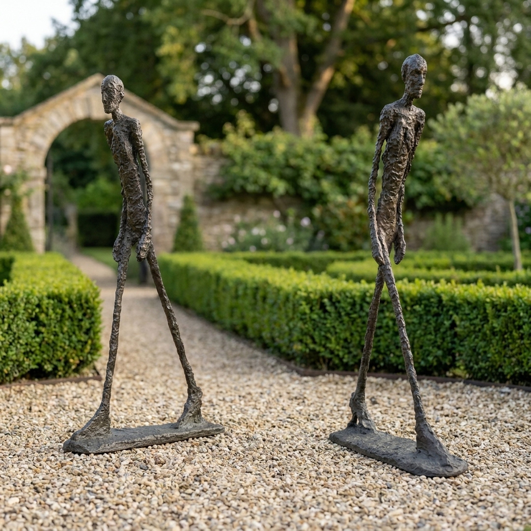 Two Large Modern Elongated Walking Male Figure Bronze Sculptures by Giant Sculptures on a gravel garden path, dark textured patina, facing one another near hedges.