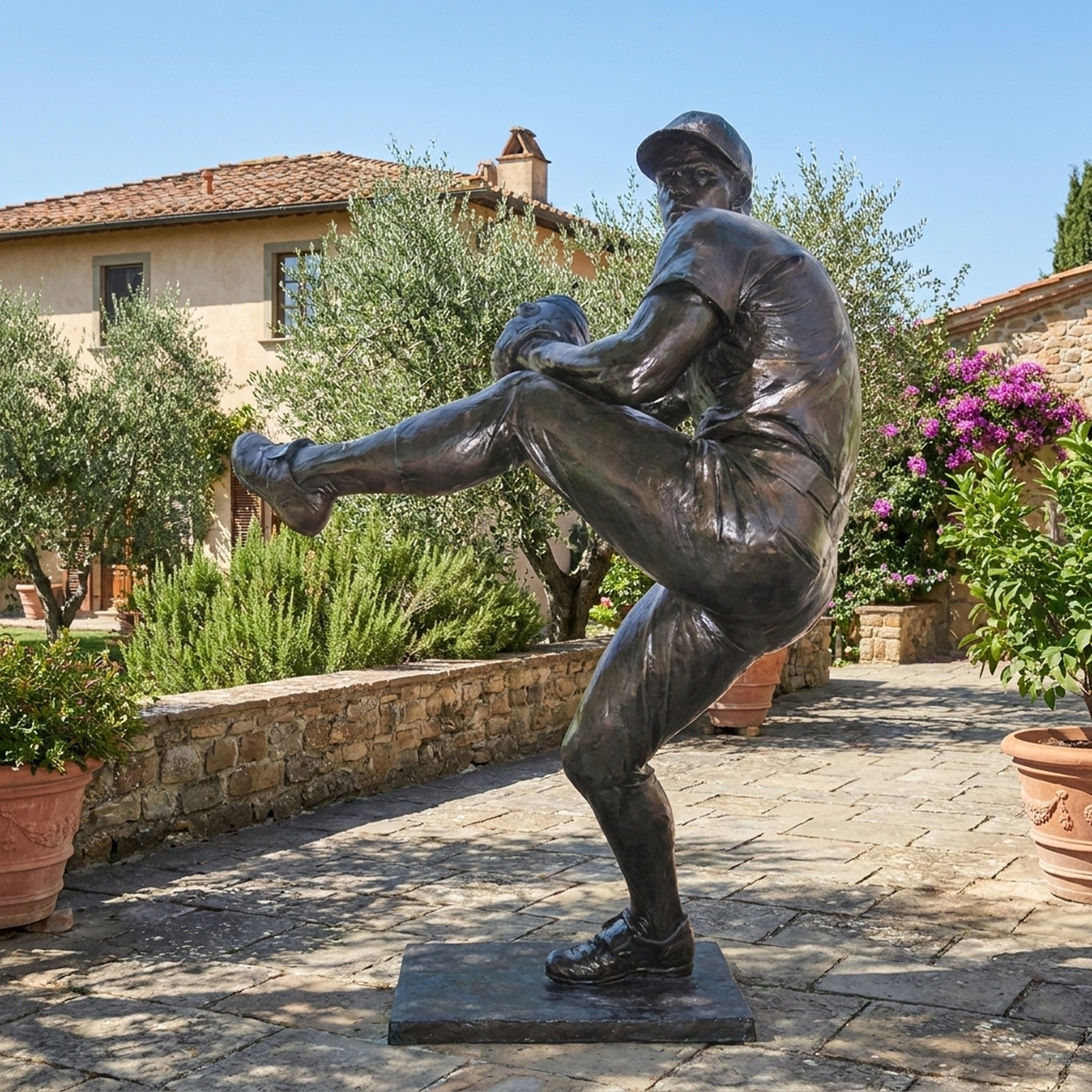 Life-Size Baseball Pitcher Bronze Statue by Giant Sculptures with dark patina, shown mid wind-up on a stone patio beside a Tuscan villa with olive trees.
