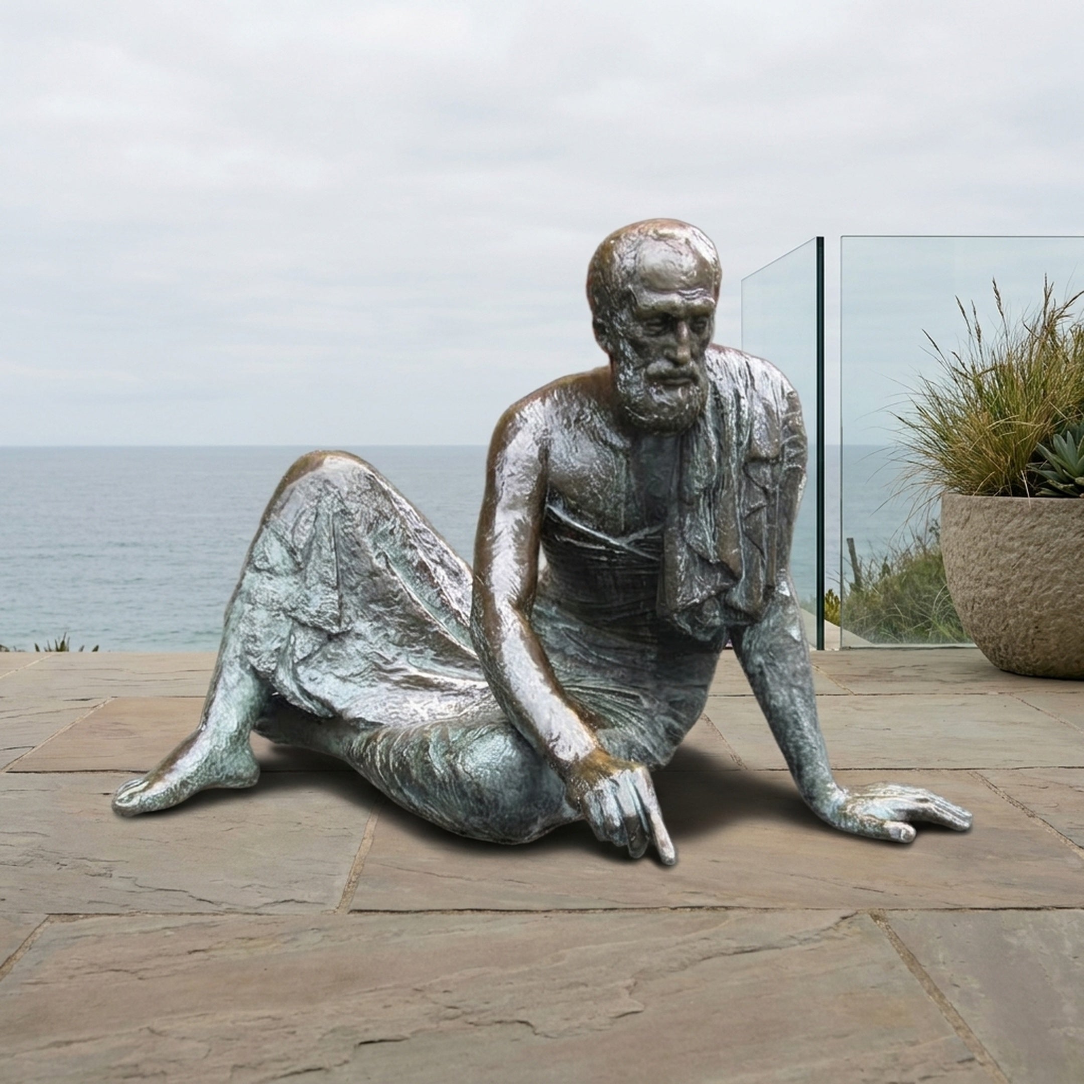 Life-Size Classical Reclining Bearded Scholar Bronze Sculpture by Giant Sculptures, shown on stone paving by a glass railing with the ocean behind.